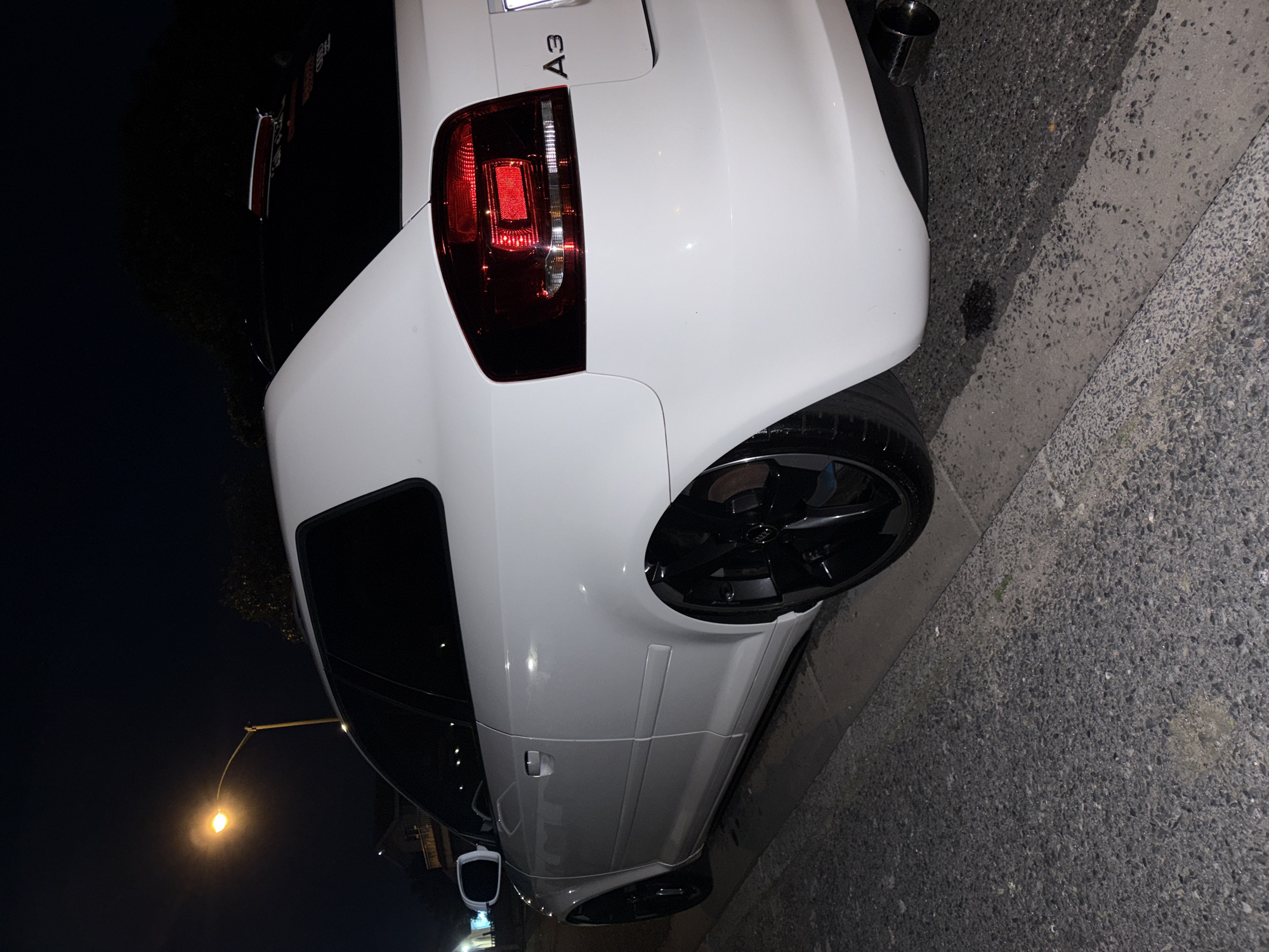 A white Audi A3 hatchback is parked at the curb of a street at night. The car is viewed from the rear driver's side quarter, with the rear wheel and part of the driver's side door visible. The car has black rims and appears to be lowered. The street is made of asphalt and there is a concrete curb. A street light illuminates the scene from the upper left. In the background, trees are visible. The car has a sticker on the rear window that appears to be red and white. The license plate is not clearly visible. The scene is dark and typical of a residential street at night. It is not possible to determine the specific location within Goodwood, South Africa based on the provided image.