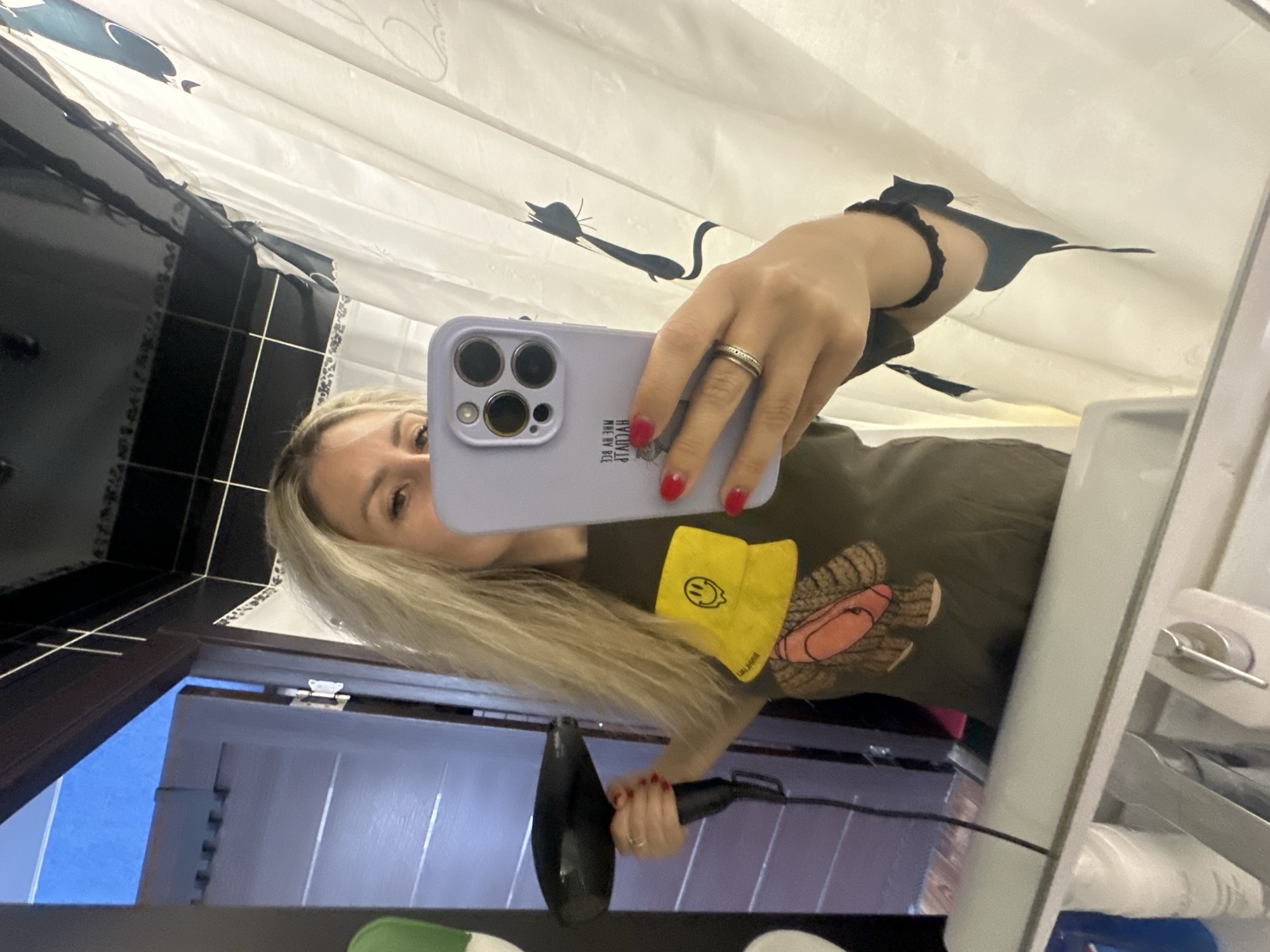 A woman with blonde hair is taking a selfie in a bathroom mirror. She is holding a black hairdryer in her right hand and her phone in her left hand, which is a light purple color. She is wearing a dark t-shirt with a graphic print of a teddy bear wearing a yellow bucket hat with a smiley face. The bathroom has a white shower curtain with black cat silhouettes on it and a black tiled ceiling. A doorway with a white paneled door is visible in the reflection of the mirror. There are bottles and a faucet visible in the foreground of the mirror's reflection. The image appears to be taken in Novoaltaysk, Russia, based on the location tag.