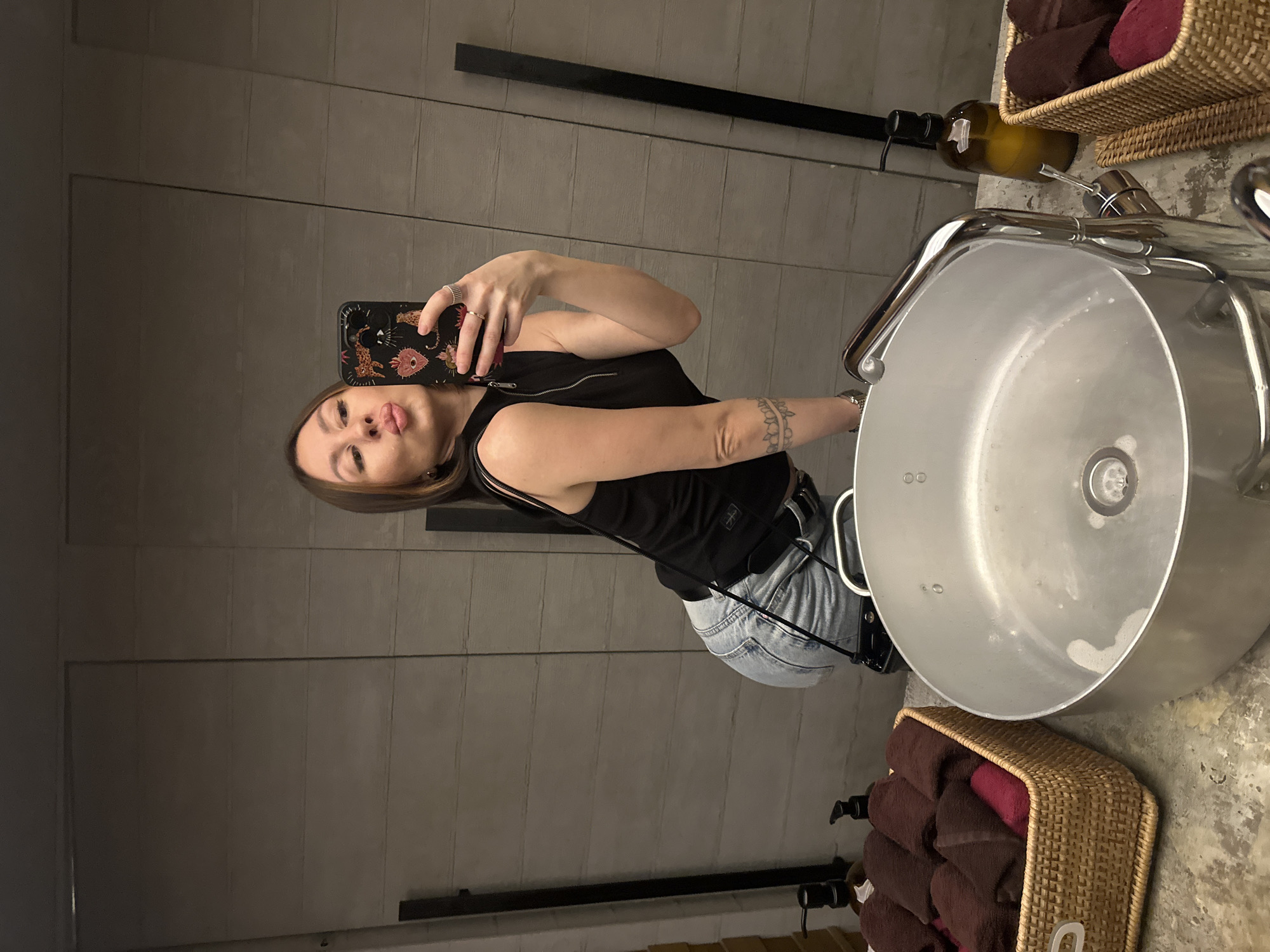 A woman is taking a selfie in a bathroom. She is wearing a black tank top and light blue jeans. The bathroom has a modern sink with a chrome faucet. There are baskets of towels on either side of the sink. The walls are gray and there are black shelves on the wall. There is no indication of Izhevsk, Russia.