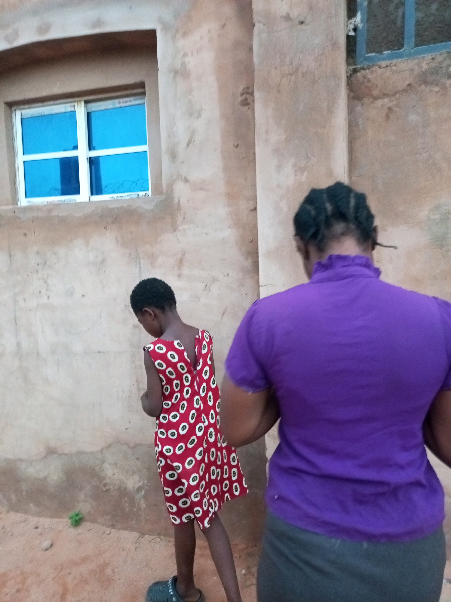 The image depicts an outdoor scene in Tempure, Nigeria, where two individuals are present in front of a building with a textured, earthen-toned wall. A window with bright blue glass and a white frame is visible on the wall to the left. The ground is composed of reddish-brown dirt and sand.
In the foreground, a person wearing a bright purple shirt and a dark skirt is facing away from the camera, with their back towards the viewer. Their hair is styled in braided cornrows. To the left of this individual, a child is also facing away from the camera, wearing a red dress patterned with white circular designs outlined in black. The child is wearing dark footwear. The child is positioned slightly in front of and to the left of the person in the purple shirt. The interaction between the two individuals is unclear as they are both facing away. The overall lighting suggests it is daytime.