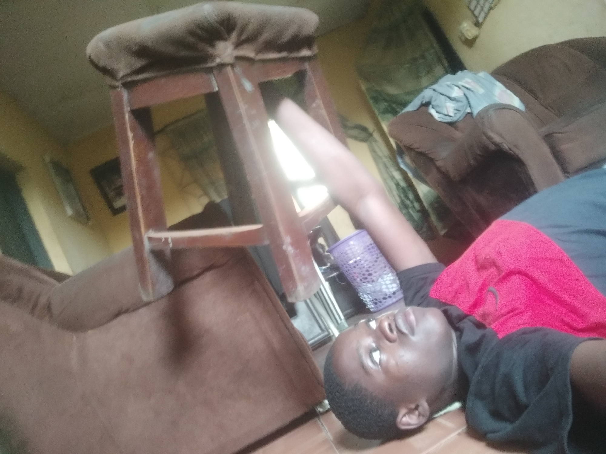 A person is lying on their back on a tiled floor indoors, with a wooden stool balanced above them. The person is wearing a black shirt and a red and gray shirt underneath. A brown sofa is to the left of the person, and another brown sofa with a blue and white cloth draped on it is to the right. A purple plastic basket sits near the person's head. A picture frame is on the wall in the background. The location is Unknown, Unknown.