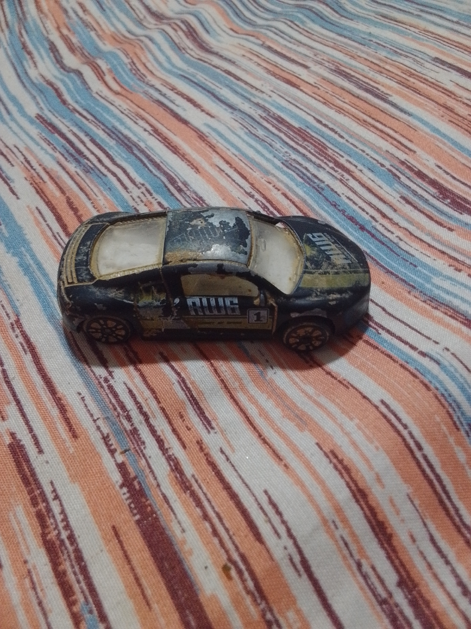 A close-up, high-angle shot shows a weathered black toy race car with gold and white racing stripes, and the number "1" on its side, resting on a striped fabric surface. The fabric is a mix of white, coral orange, and dark red-brown stripes that run diagonally from the top left to the bottom right of the image. The car appears to be made of metal and plastic, with visible wear and tear, including scuffs and peeling paint. The windows are a translucent white, and the wheels are black with a detailed spoke design. The car is oriented with its front facing towards the right side of the frame and its rear towards the left. The lighting is overhead and appears to be natural, casting soft shadows. There are no people or other significant objects visible in the frame. The scene is static and does not depict any action.