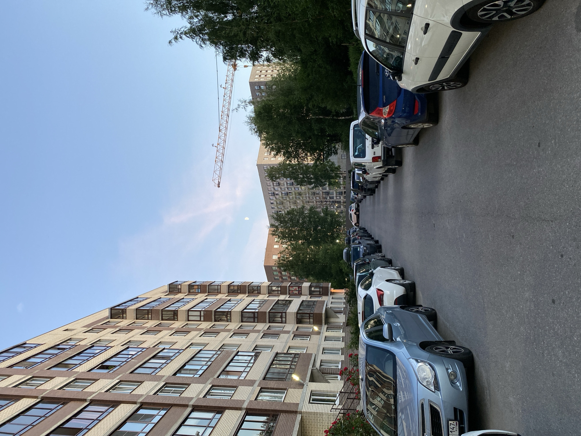 A residential street in Kanisty, Russia, is lined with parked cars and apartment buildings. The buildings, constructed with brick and concrete, appear to be modern and tall, featuring numerous windows and balconies. A construction crane is visible in the distance, suggesting ongoing development in the area. The sky is clear with a hint of pale moonlight visible. The street is paved and shows signs of wear. No individuals are present in the scene. The overall impression is one of a typical urban residential environment.