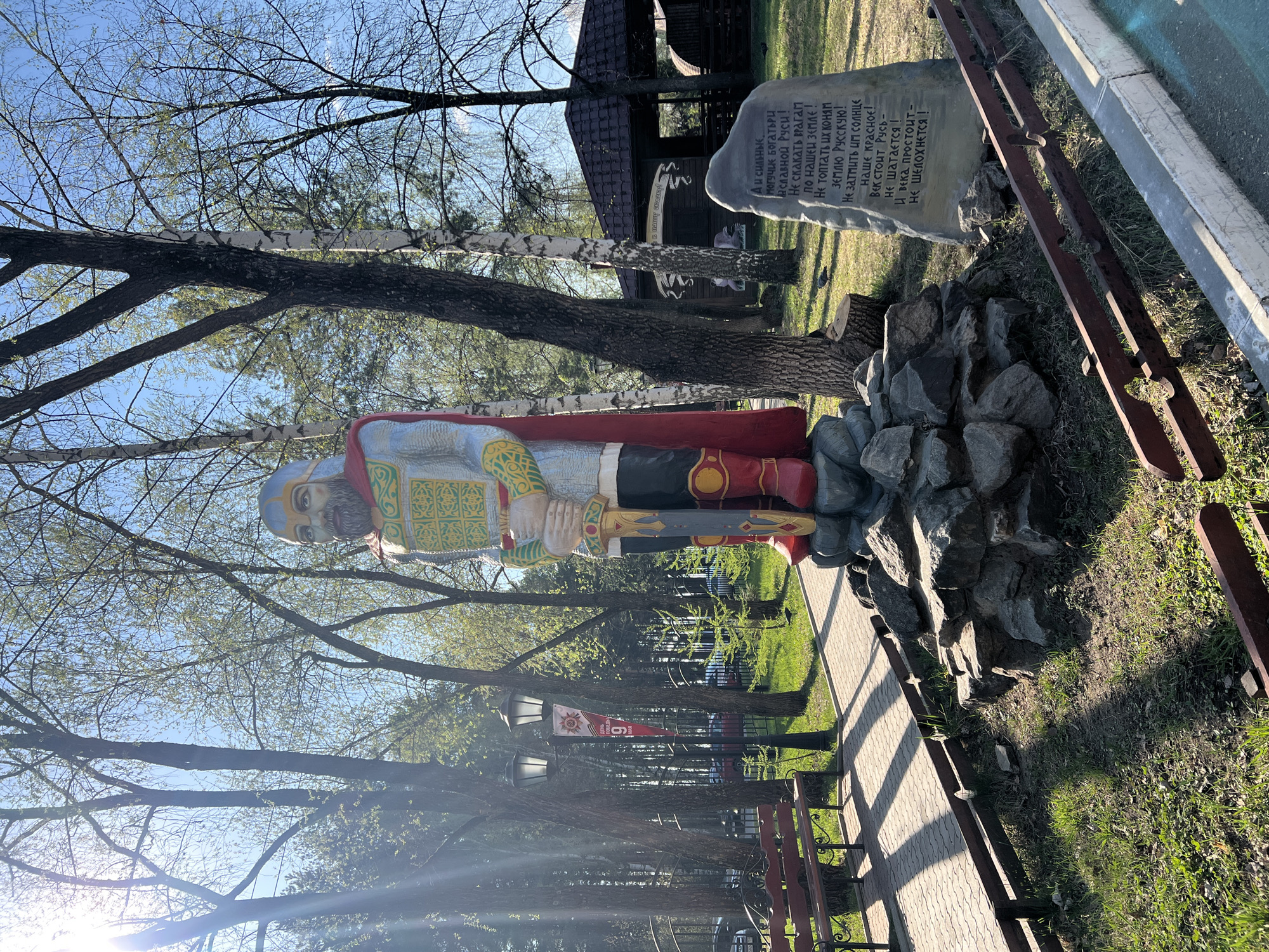 The image shows a statue of a Russian knight or warrior in a park in Chaikovsky, Russia. The statue is standing on a base of rocks and is located next to a stone plaque with Russian text. In the background, there are trees, a gazebo, and a fence. Banners on lampposts in the background seem to commemorate Victory Day (May 9). The pathway is paved with interlocking stones.