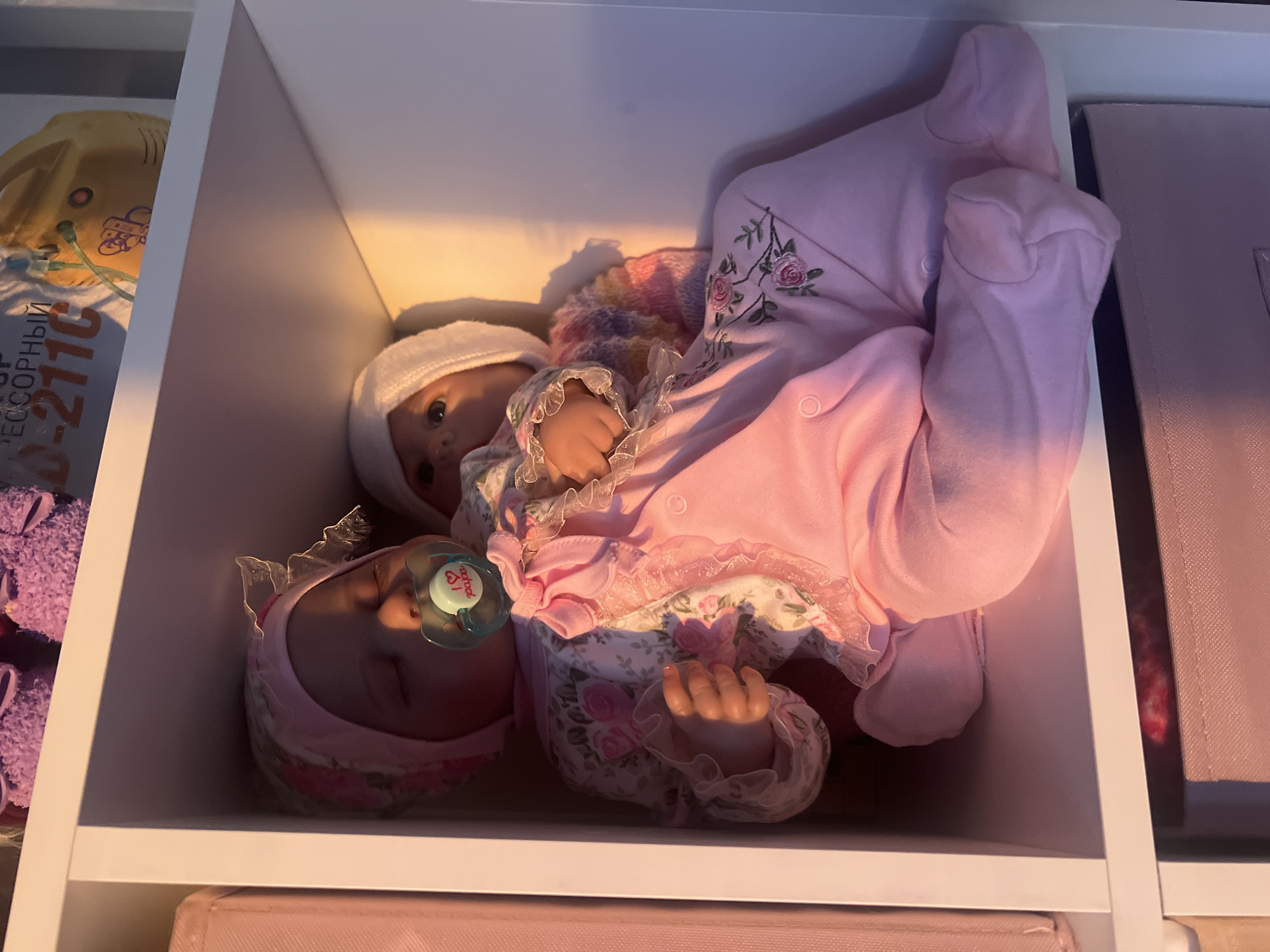 Two dolls are placed in a white storage cube. One doll is wearing a pink floral dress with lace trim, a pink bonnet, and has a pacifier in its mouth. The second doll is wearing a white cap, a colorful sweater with a frilly collar, and a pink onesie with floral embroidery on the sleeve. The storage cube is part of a larger shelf unit. In the background, other items are visible including a yellow object with Cyrillic writing, a purple object, and a pink storage box. The location is not definitively identifiable as Votkinsk, Russia.