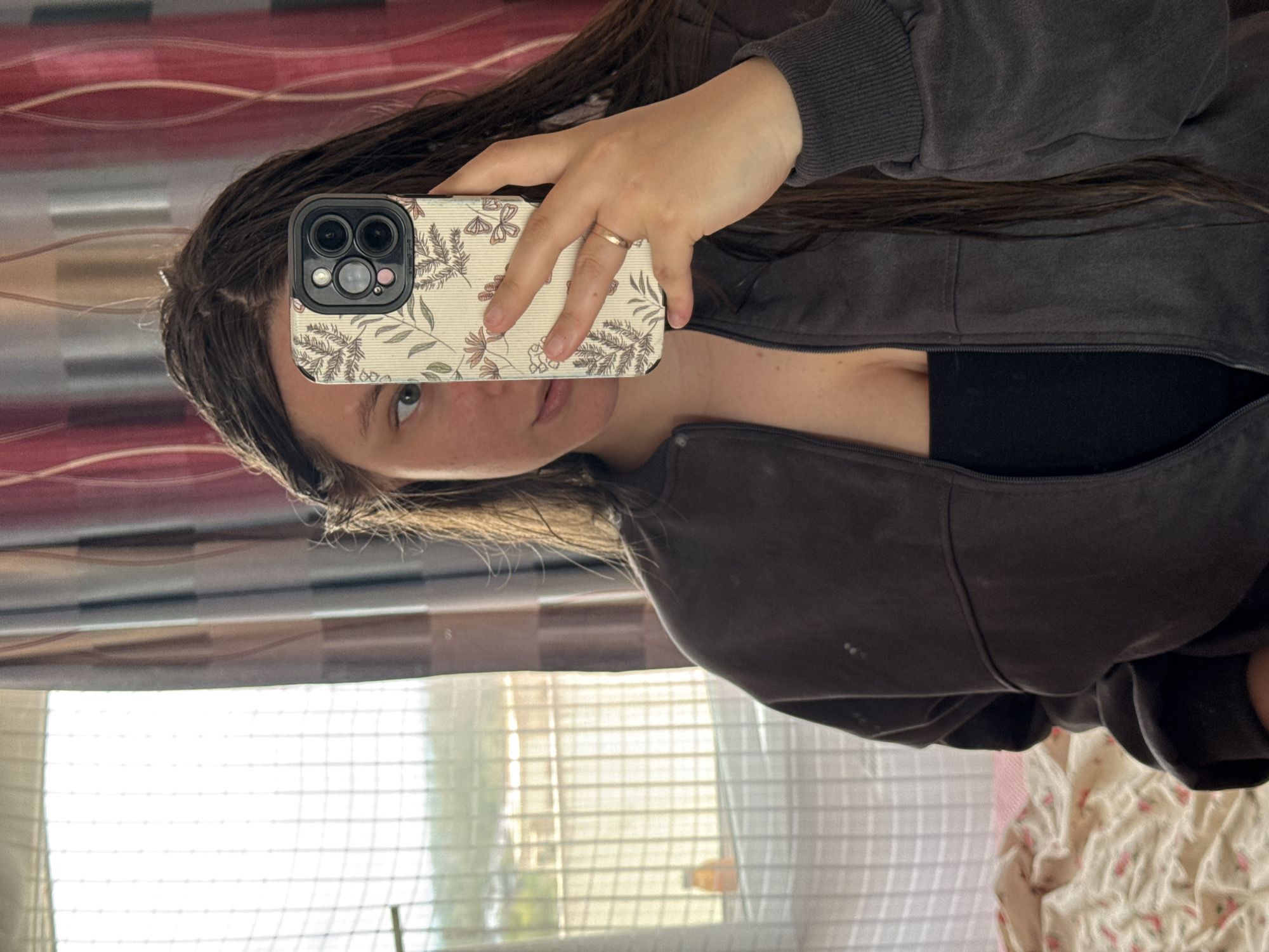 A woman takes a selfie indoors. The phone, with a floral patterned case, obscures the lower half of her face. She is wearing a dark gray zip-up jacket over a black top. A gold ring is visible on her left hand. The background shows a window with blinds and a red and white wall. The location in the city of Krasnodar, Russia cannot be determined from the provided image.