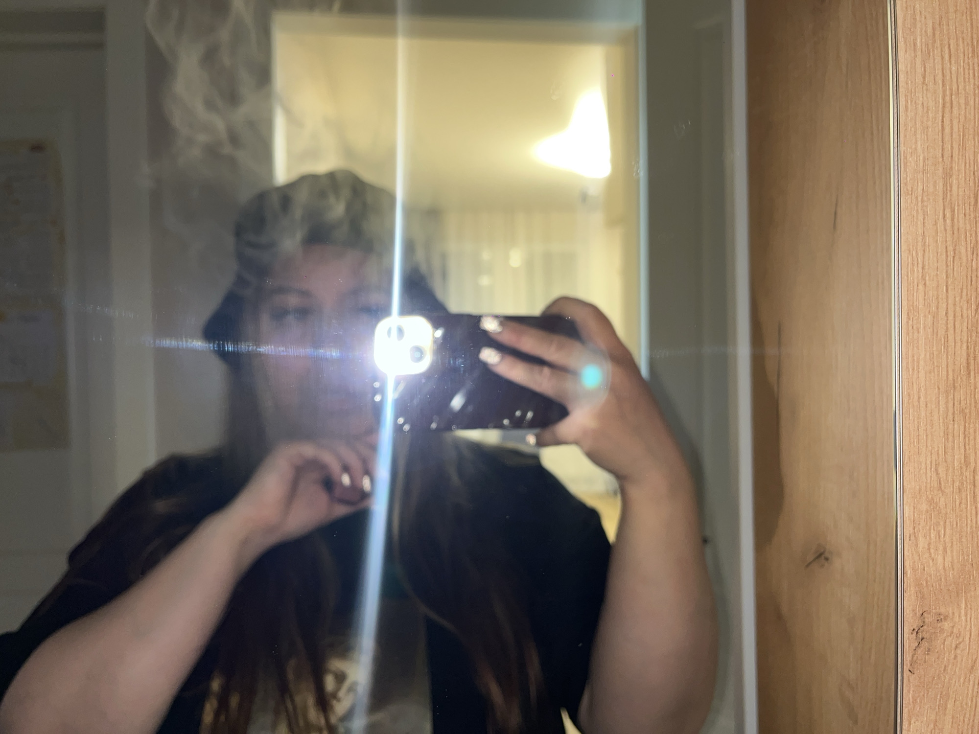 A blurred mirror selfie taken indoors in Krasnodar, Russia, shows a person holding a smartphone with a bright flash obscuring most of their face. The individual has long, dark hair and is wearing a dark top. Their hand, holding the phone, displays light-colored nail polish. The prominent flash creates a strong lens flare across the center of the image and illuminates the surrounding area.
The scene appears to be a room, possibly a bedroom or hallway, with a large mirror. Reflections in the mirror show a brightly lit interior, possibly from a ceiling light or a window, with curtains visible in the background. To the right of the mirror, a wooden wardrobe or piece of furniture is visible, displaying a natural wood grain and a vertical metal strip. The mirror surface itself shows signs of smudges or dust, and there are some visible streaks.
The strong artificial light from the phone's flash suggests the photo was taken in a dimly lit environment, possibly at night or in the evening. No specific weather conditions are discernible, and no text is visible in the image. The overall impression is an informal, personal photograph.