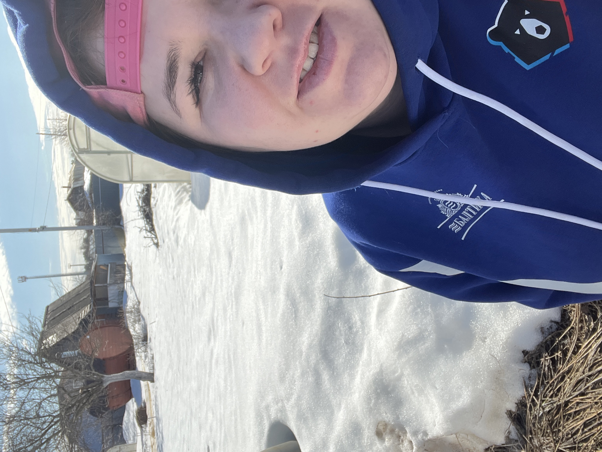 A partial frontal selfie shows a female subject outdoors in a snowy, sunny environment in Chaikovsky, Russia. The subject's face, visible from the nose to the chin, displays a slight smile with parted lips and partially exposed teeth. Her right eye is visible, featuring distinct eyelashes. She wears a blue hooded sweatshirt with white drawstrings, a partially obscured "БАЛТИКА" (Baltika) logo on the left chest, and a black bear's head logo with blue and red accents on the right chest. A pink baseball cap is worn backward under the hood. The foreground consists of snow-covered ground with patches of dry brown vegetation. The background features a bare deciduous tree, a wooden house with a steeply pitched roof and porch, two prominent red barrel-shaped structures (likely saunas) to the left of the house, and a translucent arch-shaped greenhouse to the right. Utility poles and a tall metal mast are visible in the distance under a bright blue sky.