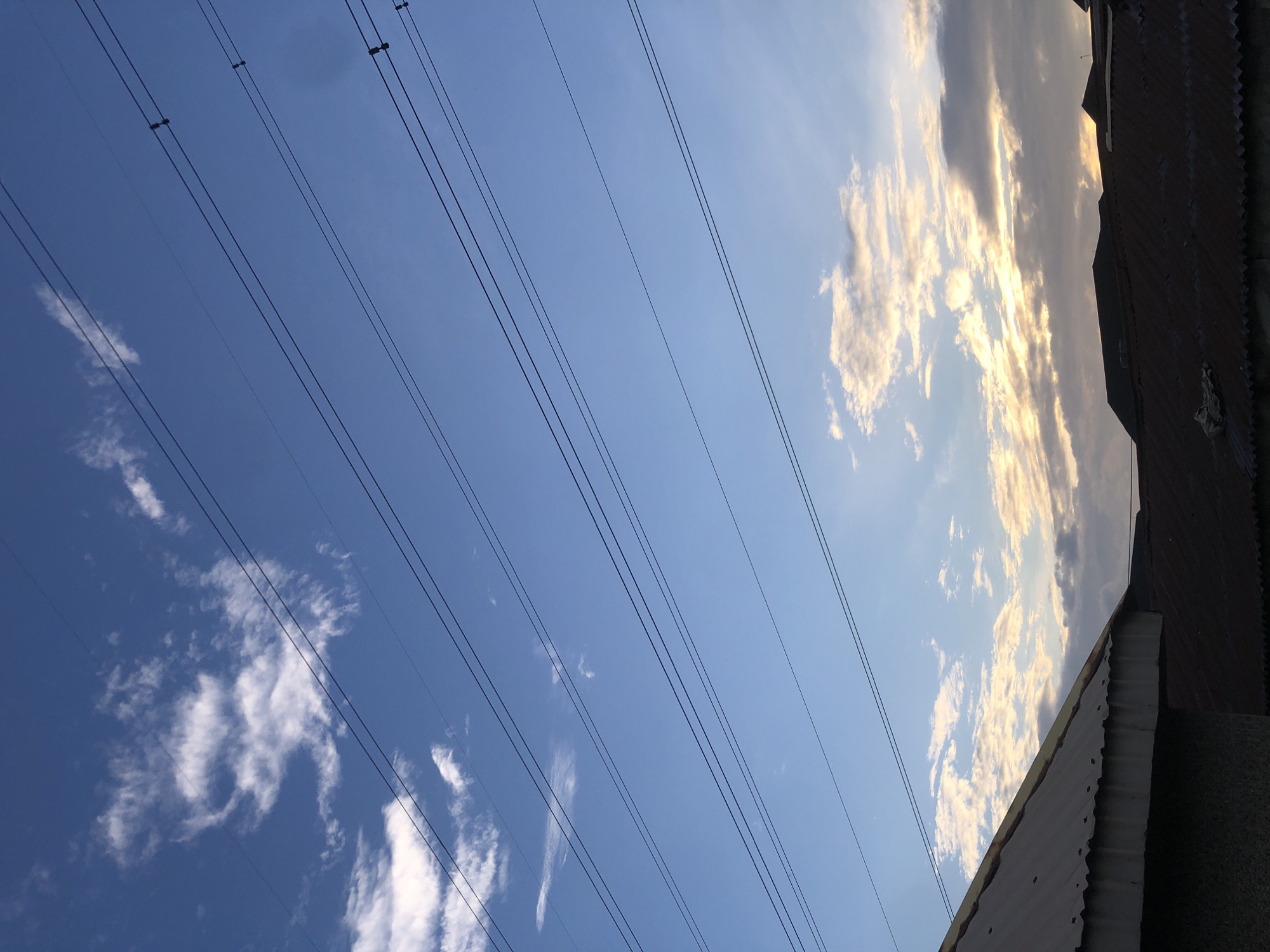 This is an outdoor, eye-level, vertical, medium shot of utility wires stretching across a blue sky at dusk over Ado, Nigeria. Several dark, parallel utility wires are strung across the upper half of the frame. In the lower portion of the image, the dark silhouettes of several corrugated metal rooftops are visible, with a lighter colored, slightly weathered rooftop in the foreground on the left. The sky is a clear blue transitioning to a lighter hue near the horizon, where the setting sun is partially obscured by scattered, wispy clouds. Some of these clouds are illuminated with a warm, golden light from the sun. No people or animals are present in the scene.