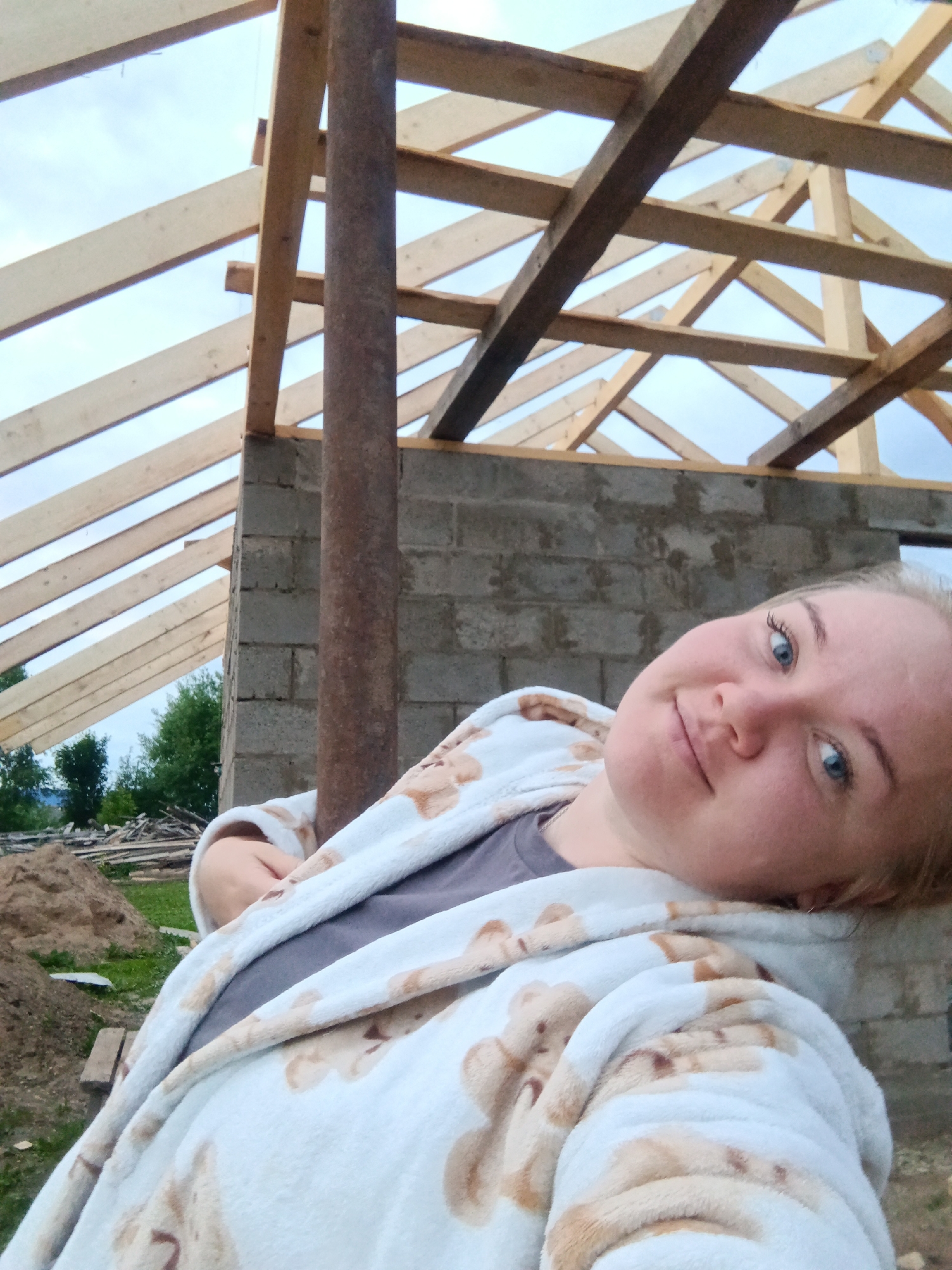 A young woman with blonde hair and blue eyes is in the foreground, looking at the camera with a slight smile. She is wearing a white robe with a teddy bear pattern and a gray t-shirt underneath. Her head is tilted to the side, resting on her shoulder. Behind her, a construction site is visible, with the wooden frame of a roof under construction and concrete block walls. A rusty metal pole stands vertically between the woman and the wall. In the background, there are piles of lumber and trees. The scene appears to be outdoors, with a cloudy sky overhead. The location is implied to be in the city of Fokinskoye, Russia, given the context of the prompt.