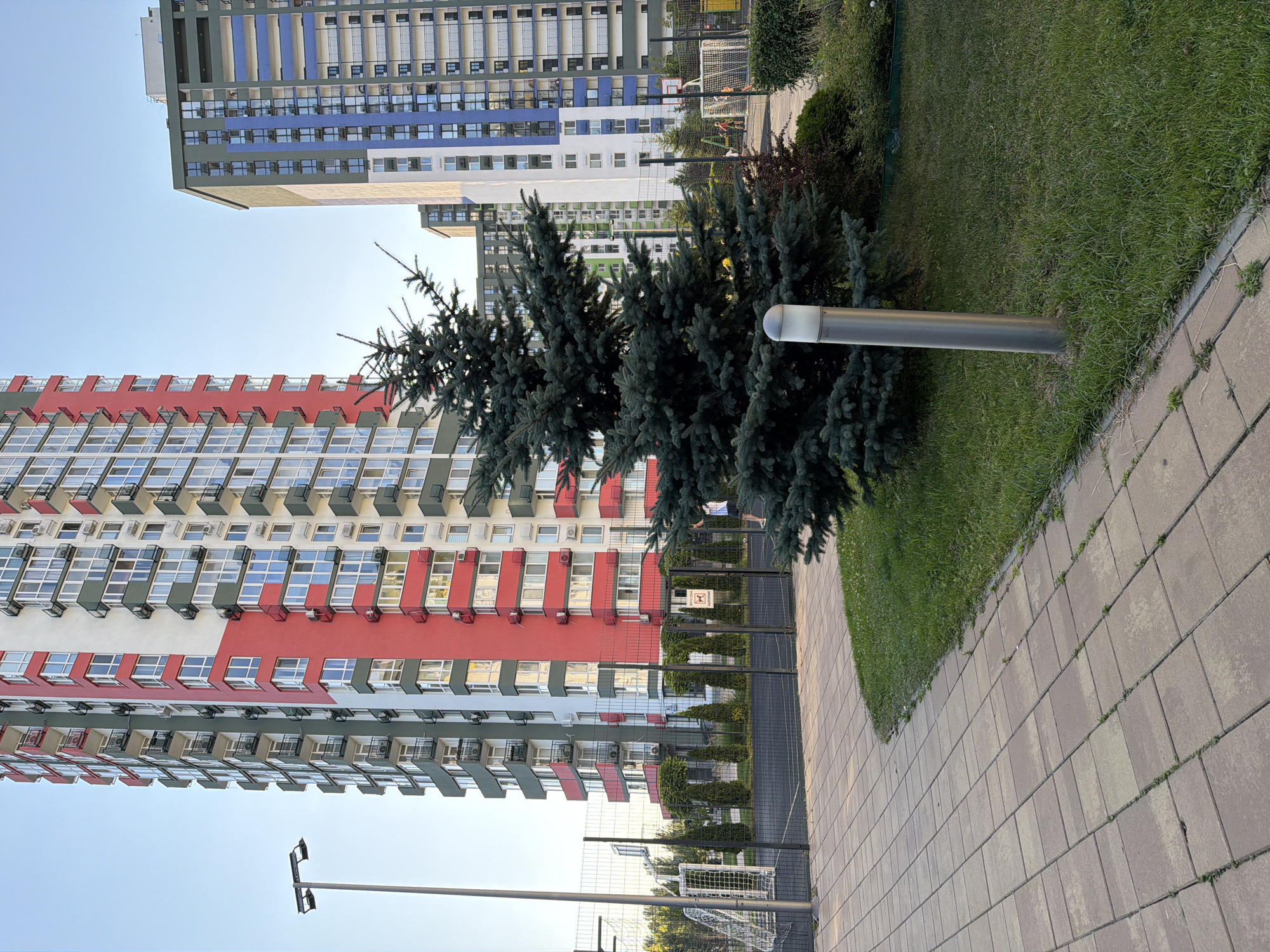 In Kyiv, Ukraine, a multi-story residential building with a prominent red and green facade stands tall against a clear sky. In the foreground, a paved pathway leads towards a fenced sports court, which features a soccer goal. A mature coniferous tree is positioned between the pathway and the court. Beyond the immediate sports area, another taller building with a blue and grey facade is visible, suggesting a modern residential complex. The scene appears to be set in a public or communal outdoor space within a city environment.