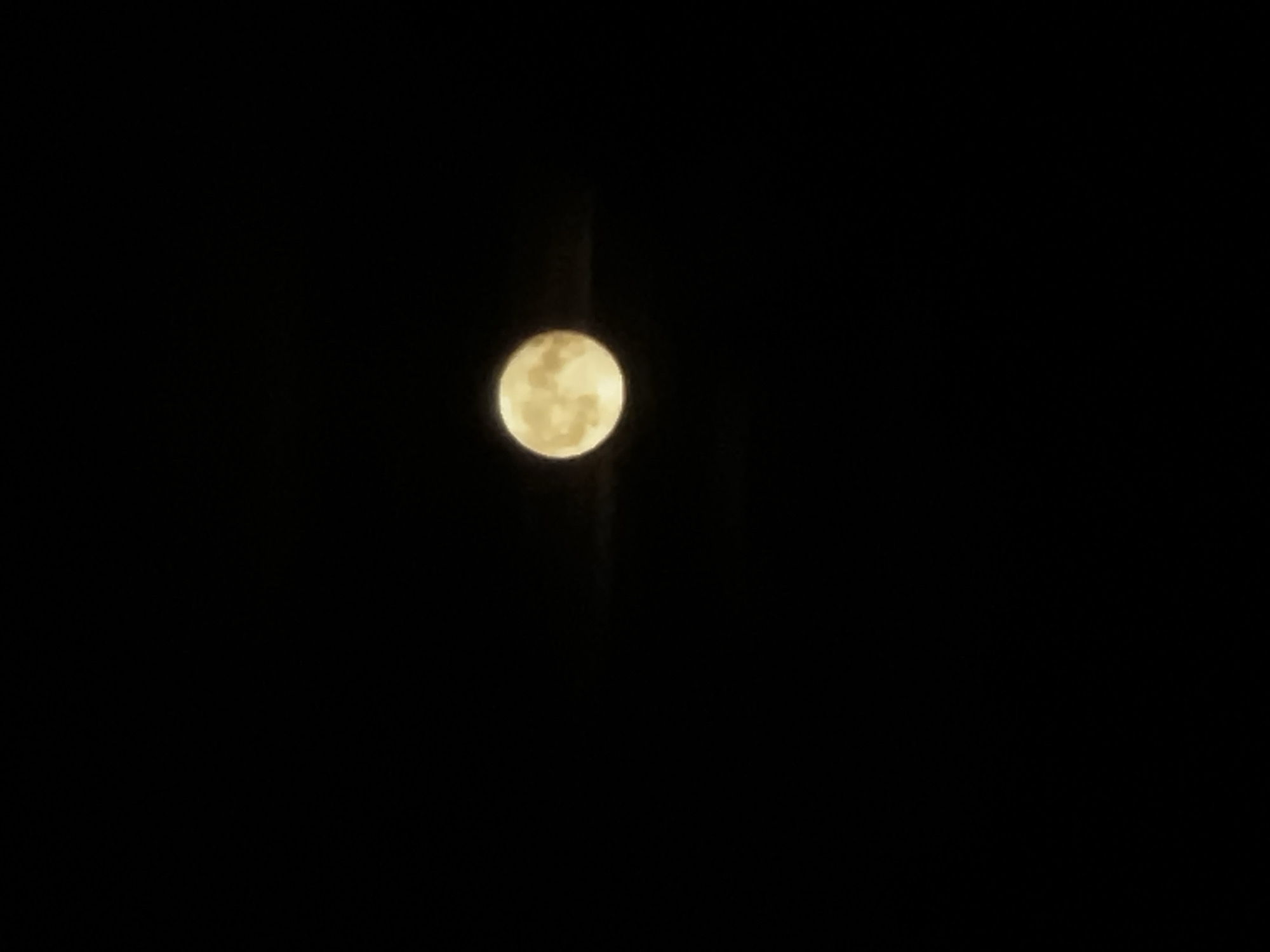 The image displays a full, circular moon, prominent and bright, against an entirely dark night sky. The moon exhibits a warm, yellowish hue, with subtle variations in its surface texture suggesting lunar features, though these details are slightly soft or blurred. A faint, narrow vertical light streak is subtly visible, extending directly above and below the moon, possibly an effect of light diffraction or atmospheric conditions. The surrounding environment is completely black, indicating a clear, cloudless night. No other celestial bodies, landscapes, or artificial lights are visible in the frame. The photograph captures the moon in isolation, as observed from Khimki, Russia. There is no visible text in the image.