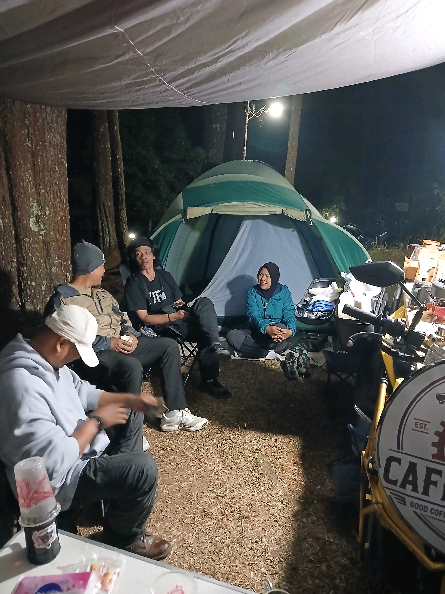 The image depicts a camping scene at night in Berjo, Indonesia. Four individuals are present. Three men are seated in camping chairs, engaged in conversation. One man in a grey beanie and tan jacket holds a white cup. Another man in a black t-shirt and black pants looks towards the left. The third man, wearing a light blue hoodie and white cap, is partially visible in the foreground. A woman in a blue jacket and headscarf is seated on the ground near a green and grey tent. Various camping equipment, including bags, shoes, a motorcycle, and a table with cups and snacks, are visible. The environment is a wooded area with tall trees, and the ground is covered in dry grass and dirt. A tarp is stretched overhead, providing shelter.
