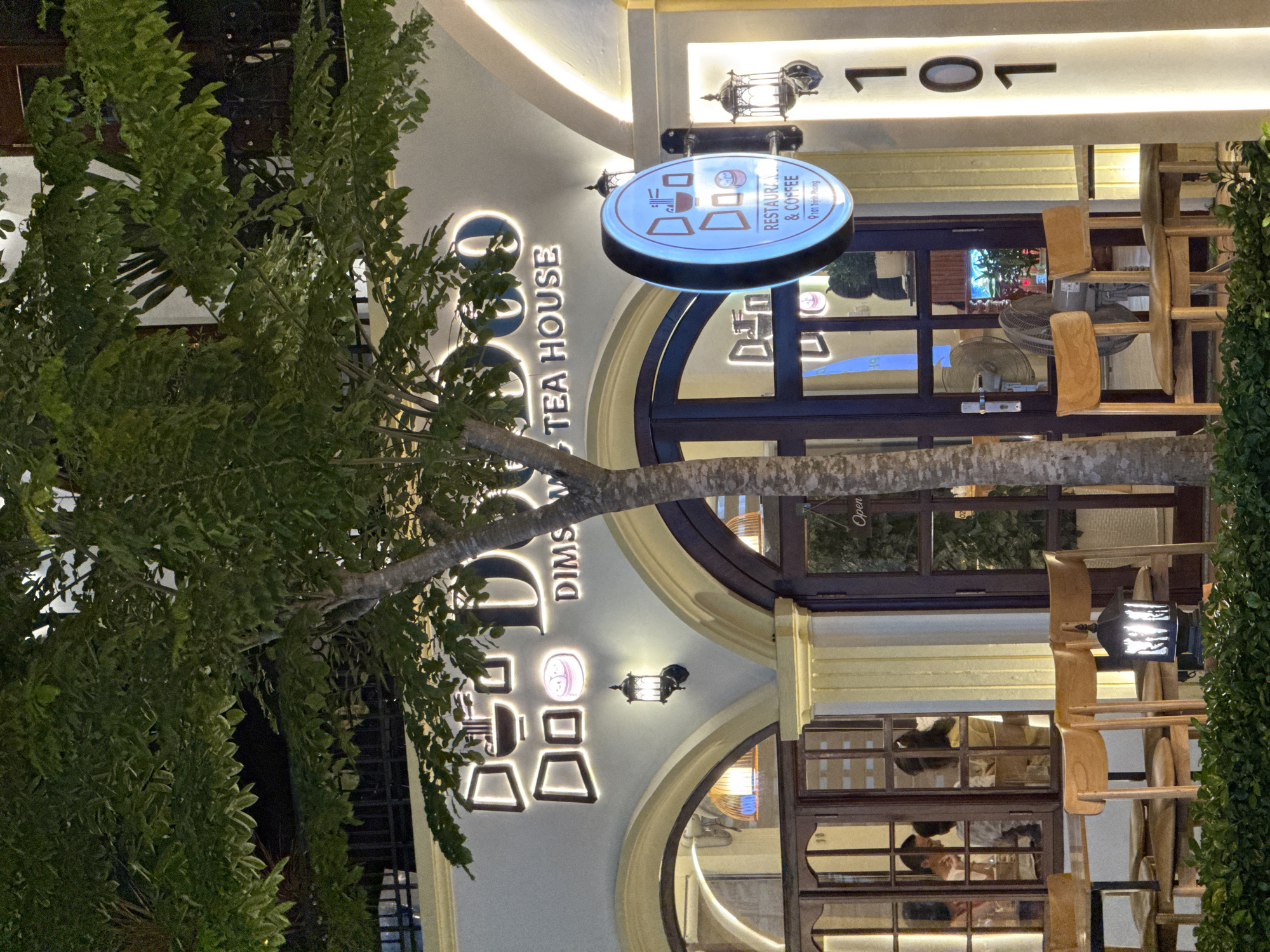 The image depicts the exterior of "Doo Doo Dim Sum Tea House" located at 101 Trinh Phong street in Nha Trang, Vietnam. The establishment features a light-colored facade with prominent neon signage that spells out "Doo Doo" and "DIM SUM TEA HOUSE". The entrance is marked by large, arched doorways with dark wood frames and glass panels. Several wooden chairs are arranged at tables outside the establishment, with some patrons visible inside through the windows. A circular illuminated sign for "Doo Doo Restaurant & Coffee" hangs from the building. A tree with lush green foliage stands in front of the entrance, partially obscuring the building's signage. The scene is illuminated by the building's lights and streetlights, suggesting it is nighttime. The overall impression is of a modern, inviting tea house and restaurant.