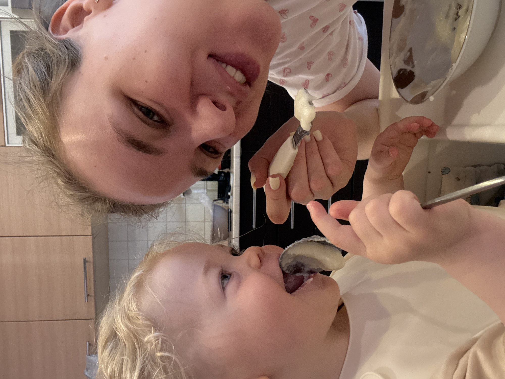 A selfie shows an adult feeding a child. The adult, appearing at the top of the image, is smiling slightly, with their face and hair angled in the opposite direction of the child. They are wearing a white shirt with a pink heart pattern and holding a small, white-handled utensil with food on it. Below, the child, with fair hair, is eating food from a spoon. A white bib is visible around the child's neck. In the background, there are wooden cabinets and a kitchen setting. The location is unknown.
