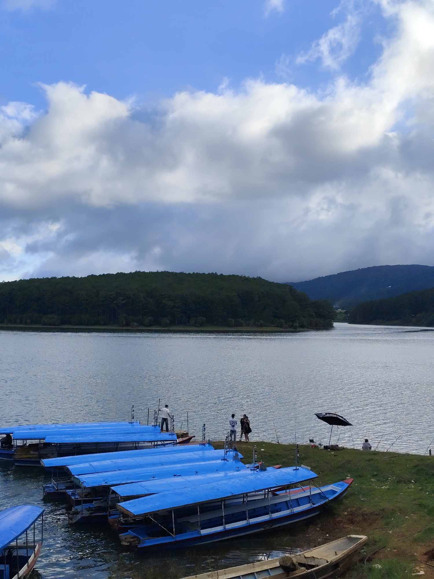 The image depicts a lake scene in Da Lat, Vietnam. Several blue-roofed boats are docked along a grassy shoreline. A group of people stand on the shore near the boats, with some holding fishing rods. An open black umbrella stands nearby, providing shade to a seated individual also fishing. The lake extends towards a densely forested peninsula. In the background, distant mountains are visible. The sky is partly cloudy with blue patches.