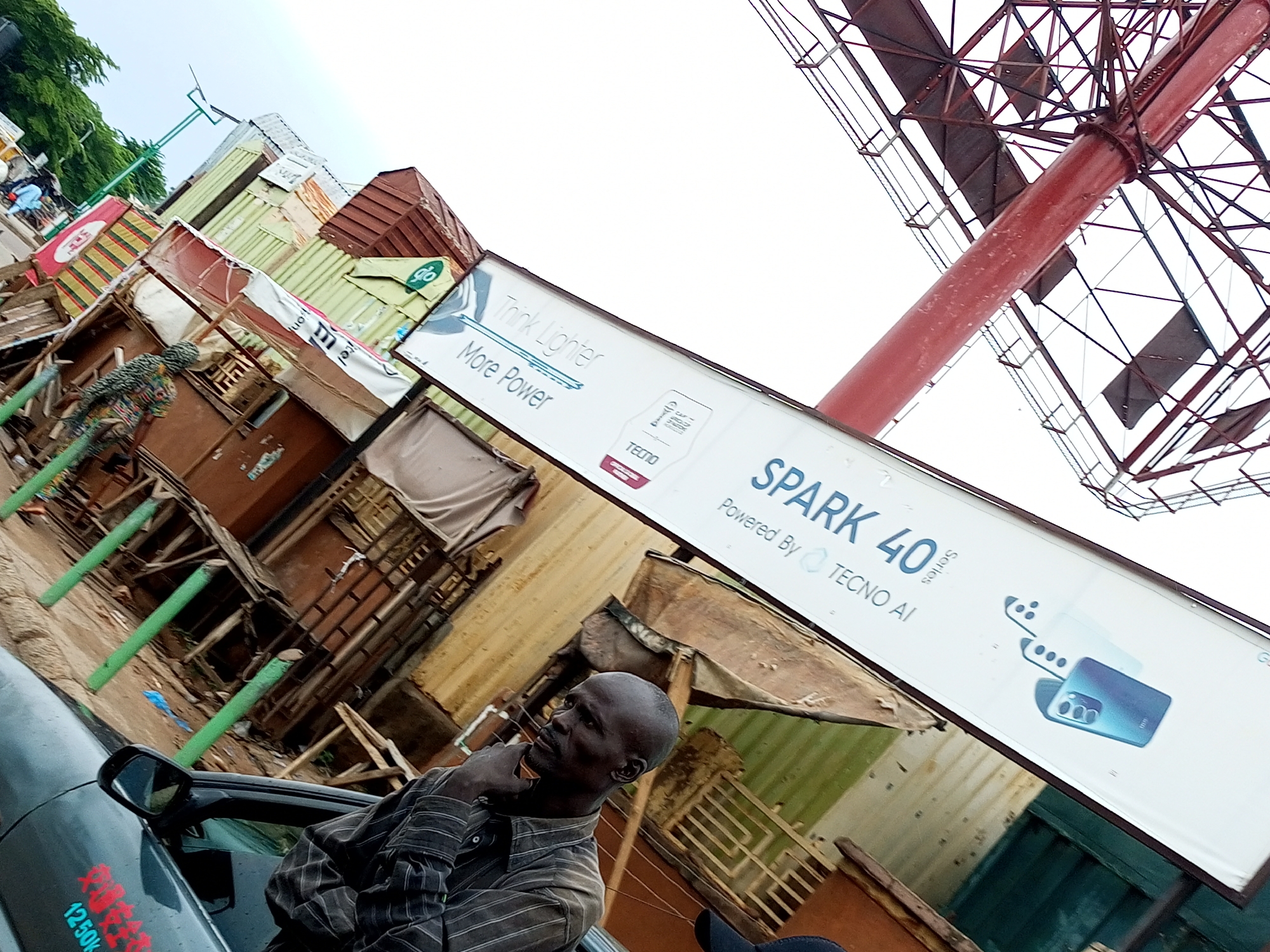 In Arawa, Nigeria, a man with a shaved head and a goatee stands next to a dark car. He is wearing a striped long-sleeved shirt and has his hand on his chin, looking thoughtfully to the left. Behind him, a street scene unfolds with various small shops and structures constructed from corrugated metal and wood. A billboard advertising "SPARK 40 Series" by TECNO AI is prominently displayed, angled upwards. Green poles line the side of the road, and the sky is overcast. The overall impression is a bustling, outdoor marketplace environment under a cloudy sky.