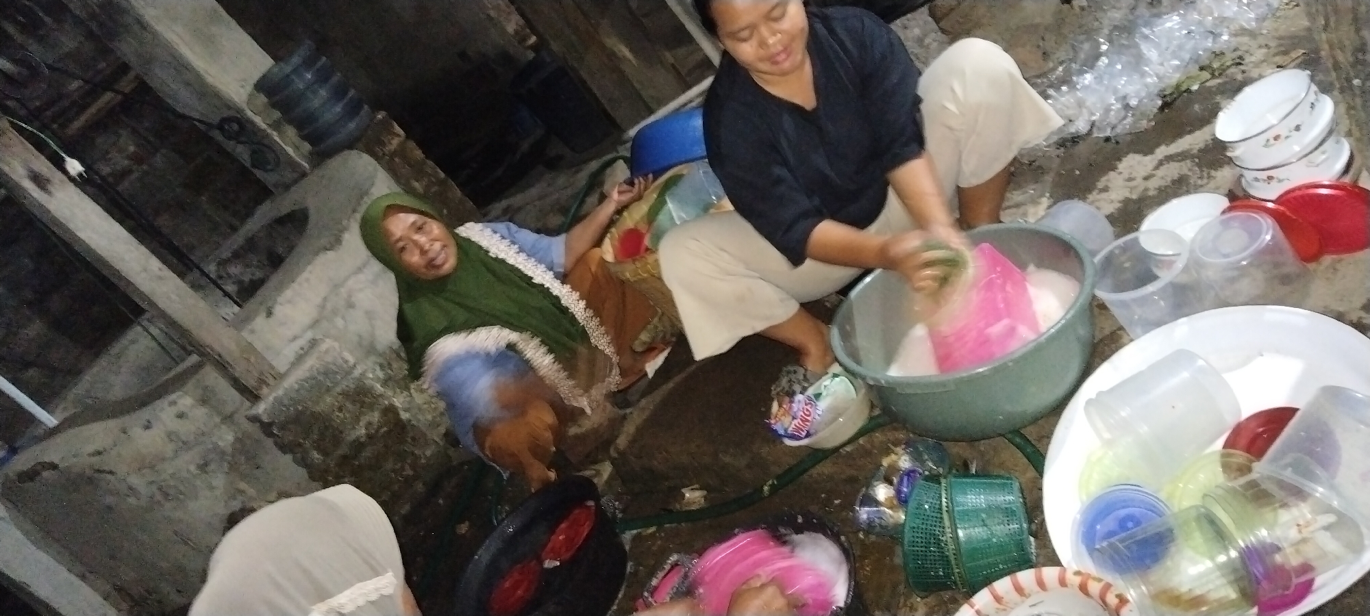 In Tanjung Ratu Ilir, Indonesia, two women are engaged in washing dishes outdoors. One woman, wearing a green hijab, is seated and appears to be resting or observing. The other woman, dressed in a black top and light-colored pants, is actively washing dishes in a large gray basin filled with soapy water. Scattered around them are various plastic containers, bowls, and a red bowl. The environment appears to be somewhat rustic, with concrete ground and wooden structures visible in the background, suggesting a potentially humble or utilitarian living space.