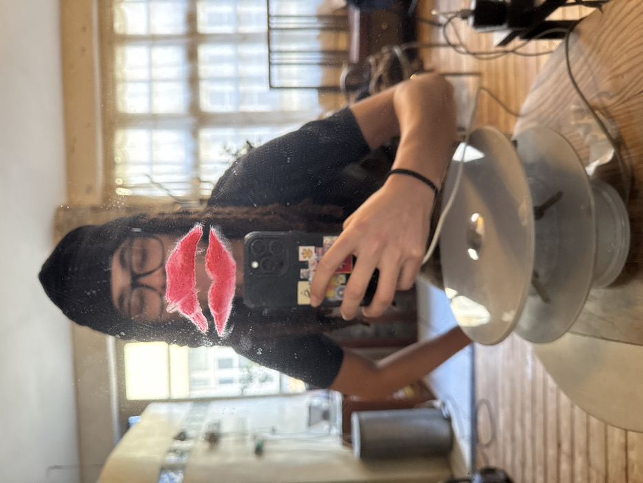 The image displays a mirror selfie captured indoors within a residence located in Paris, France. A person with dark dreadlocks, wearing a black beanie and eyeglasses, is reflected in the mirror. A prominent red lip-shaped graphic is affixed to the mirror, positioned over the reflection of the person's mouth area. The individual holds a dark-colored smartphone, adorned with multiple stickers on its back, in their left hand, which is raised to take the photograph. Their right arm is visible with a black elastic band around the wrist. They wear a dark, short-sleeved top.
In the immediate foreground, on the surface beneath the mirror, rests a multi-tiered decorative object, likely a lamp, featuring two opaque white or light grey circular plates. The mirror surface exhibits smudges and dust. The reflected background reveals an interior room with a large, multi-paned window, consistent with traditional Parisian apartment architecture, allowing natural daylight to enter. Visible elements include wooden floorboards, a light-colored wall section, and a grey cylindrical object to the left. To the right, a dark wooden furniture piece and a charging cable with an adapter are present.