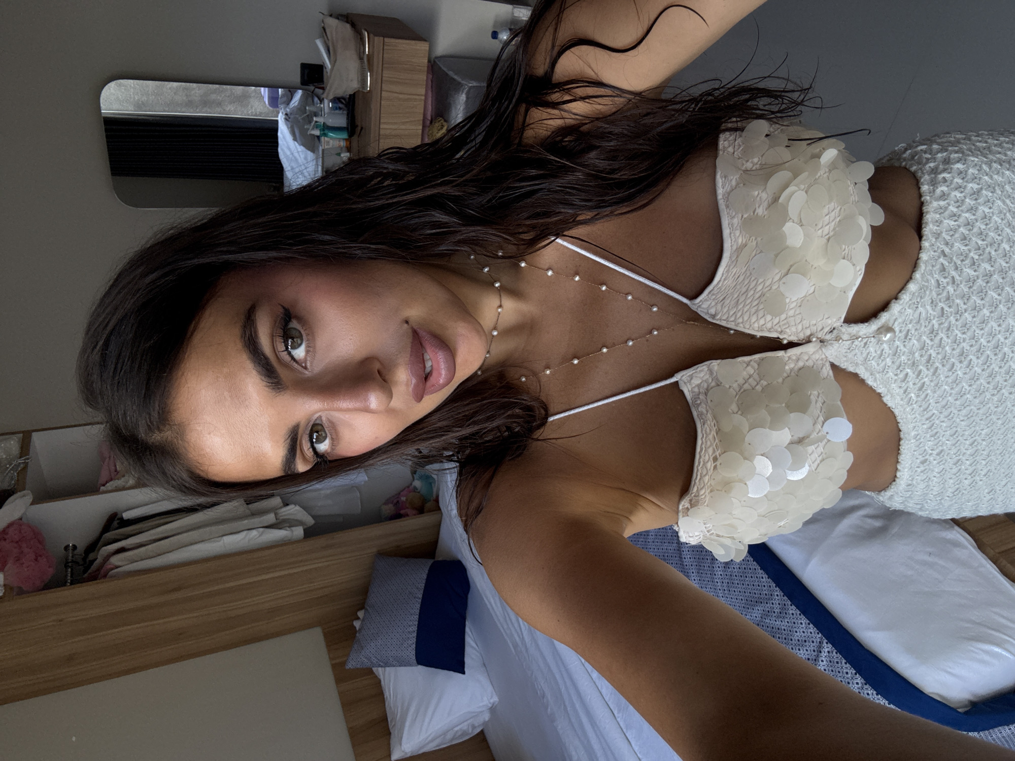 A woman is in an interior setting, likely a hotel room. She is wearing a white dress with circular, decorative appliqués on the bodice and a thin gold necklace with small pearls. Her hair is dark and appears wet, and her makeup is subtle. Behind her is a bed with a white duvet and pillows, and a blue and white decorative pillow. A light-colored wooden shelf is also visible with various personal items. Given the context, it is possible the location is in Kuta Selatan, Indonesia.