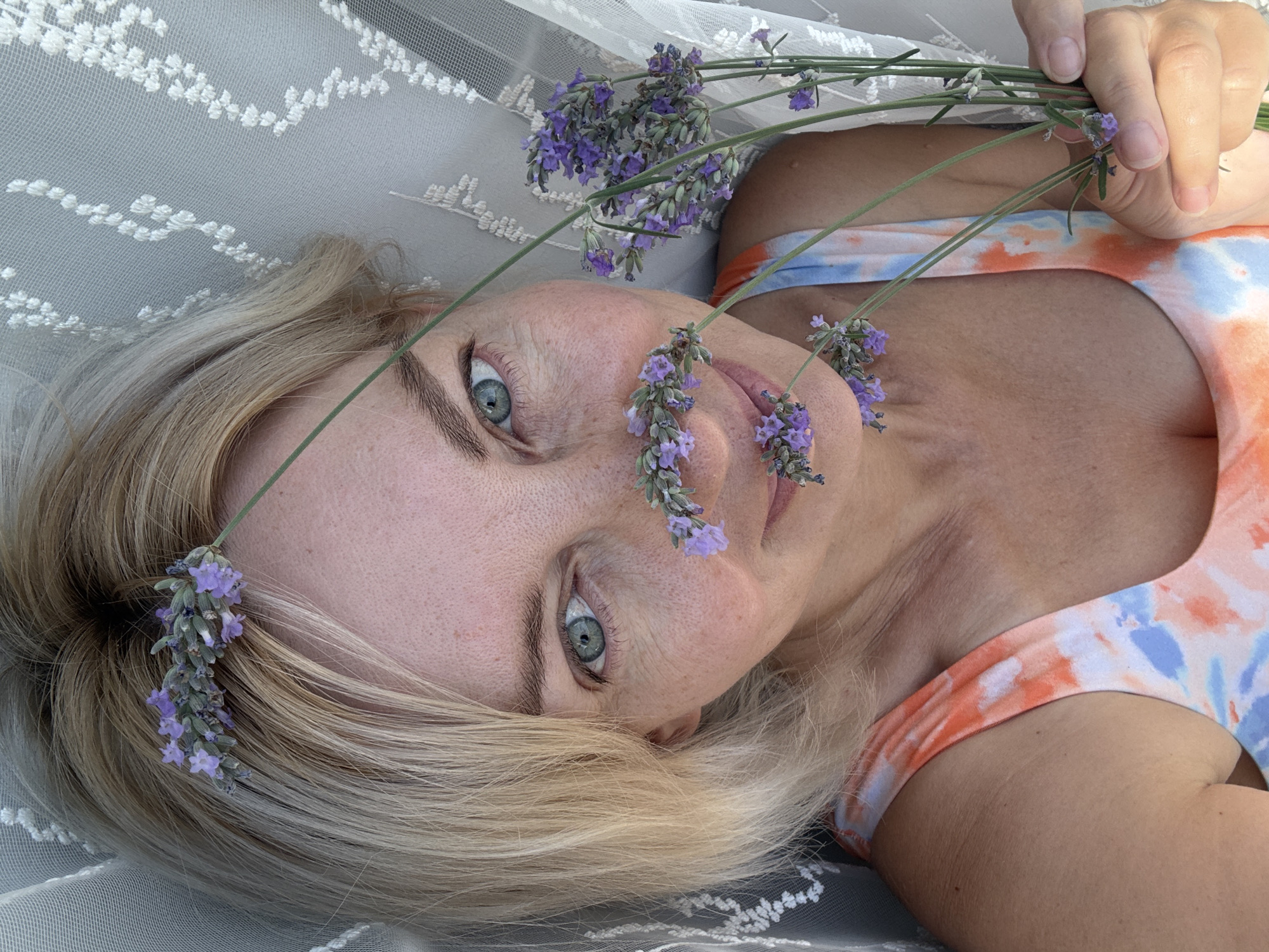 A woman with blonde hair and blue eyes lies on her back, holding sprigs of lavender. Her head is resting on a white, sheer, lacy fabric, possibly a veil or curtain. She is wearing a tie-dye tank top with orange, blue, and white patterns. Several sprigs of lavender are positioned around her face, with some resting near her nose and mouth, and one tucked behind her ear. She is holding a bunch of lavender in her right hand, which is visible in the lower right corner of the image. The background is out of focus, with the white lacy fabric being the most prominent element besides the woman and the flowers. The lighting suggests an outdoor or well-lit indoor setting. There is no discernible indication of the city of Sochi, Russia within the image itself.