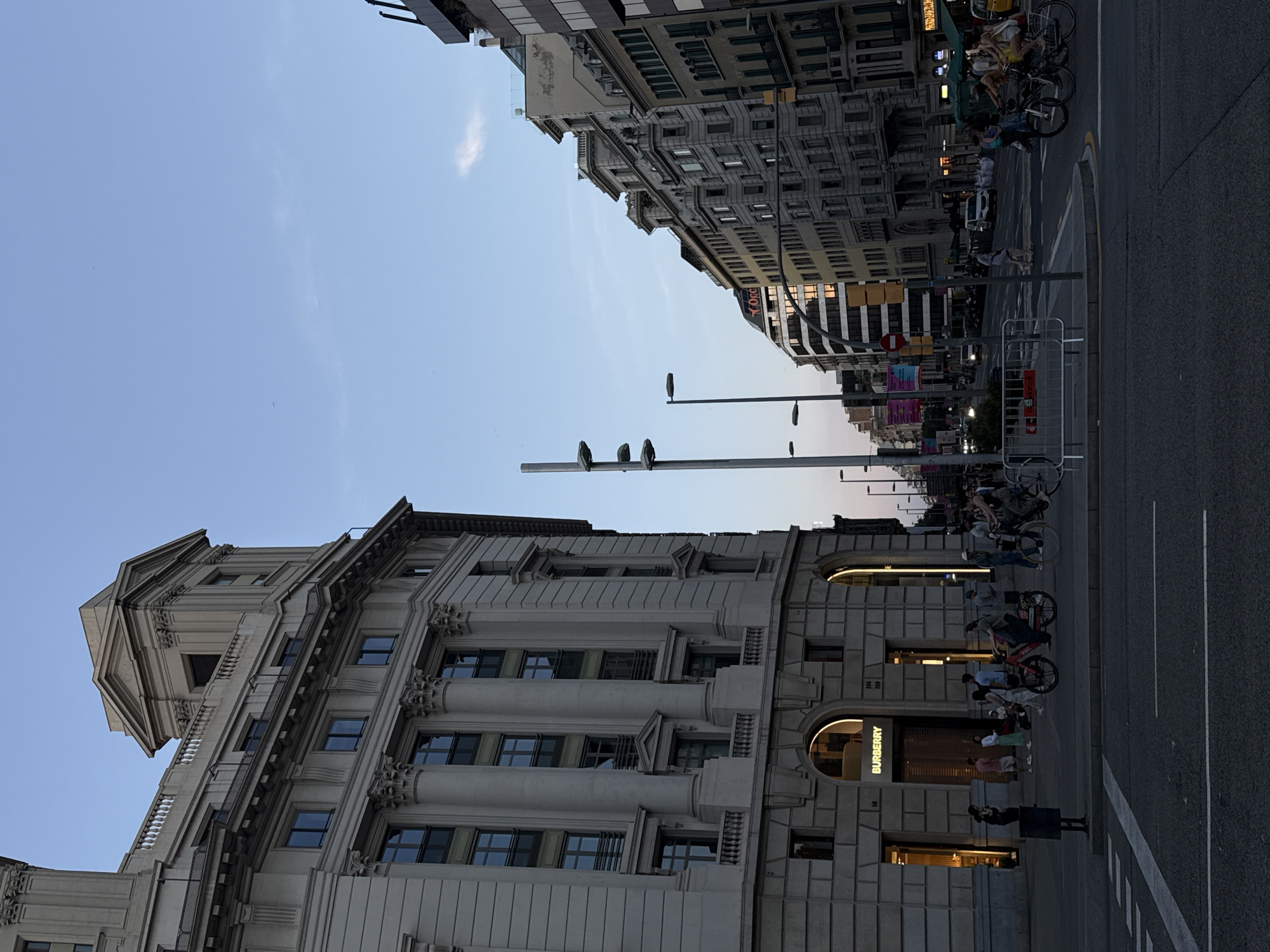 A street-level photograph captures a bustling urban scene in Barcelona, Spain, under a clear, bright blue sky with sparse wispy clouds. The image is taken from a slightly low angle, looking up at the surrounding architecture.
On the left side of the frame stands a grand, classical-style building with ornate columns, intricate cornices, and numerous rectangular windows. Its ground floor features a prominent luxury retail store, identified by the "BURBERRY" logo illuminated above a large arched display window. Several people are visible on the sidewalk in front of this building, some standing near the store entrance, others walking, and at least one person's reflection is visible in a window. A dark-haired person wearing a light-colored top and green trousers is among those near the Burberry store.
Across the street, on the right, is another multi-story building, appearing somewhat more modern with varying window styles. Further down this street, additional buildings recede into the distance. The asphalt road surface, marked with white lines, occupies the foreground and extends into the mid-ground. A tall, slender streetlight pole, topped with a horizontal arm, stands centrally in the mid-ground, with three small birds perched upon it.
The street is moderately busy with pedestrians and several bicycles parked or being used by people. Some metal barriers are set up on the sidewalk near the street. Artificial lighting from the storefronts is visible, suggesting it is late afternoon or early evening. The overall atmosphere is that of an active city street. Aside from "BURBERRY," some numbers like "216," "239," and "96" are visible on the building facade, and a partial "STOP" sign is discernible in the distance.