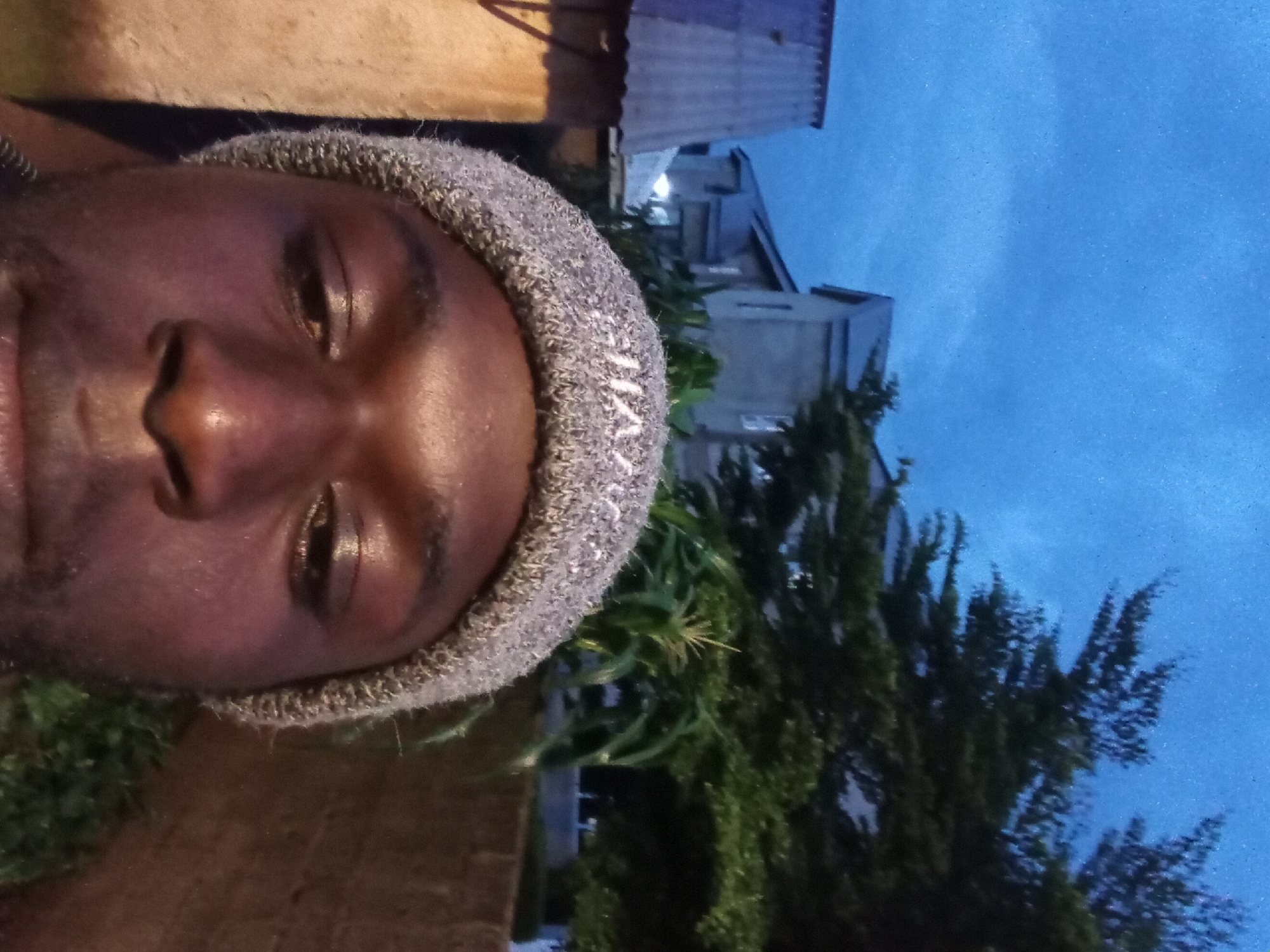 In Bukuru, Nigeria, an image captures a close-up of a man wearing a grey knit beanie. He is looking slightly down and to his left, with his right eye visible. His skin is dark, and there appears to be some sweat or moisture on his face. The background is dimly lit, suggesting it is either dawn or dusk. To the left of the man, a structure with a corrugated metal roof is visible, possibly part of a building or shed. Further behind, two-story buildings with grey facades are present, some with illuminated windows. Lush green trees, including what appears to be a corn stalk, are interspersed between the buildings. A brown brick wall is visible to the right of the man, partially obscuring more greenery. The sky above is a deep blue, with some faint cloud formations. The overall scene suggests an outdoor setting in a residential or semi-urban area.