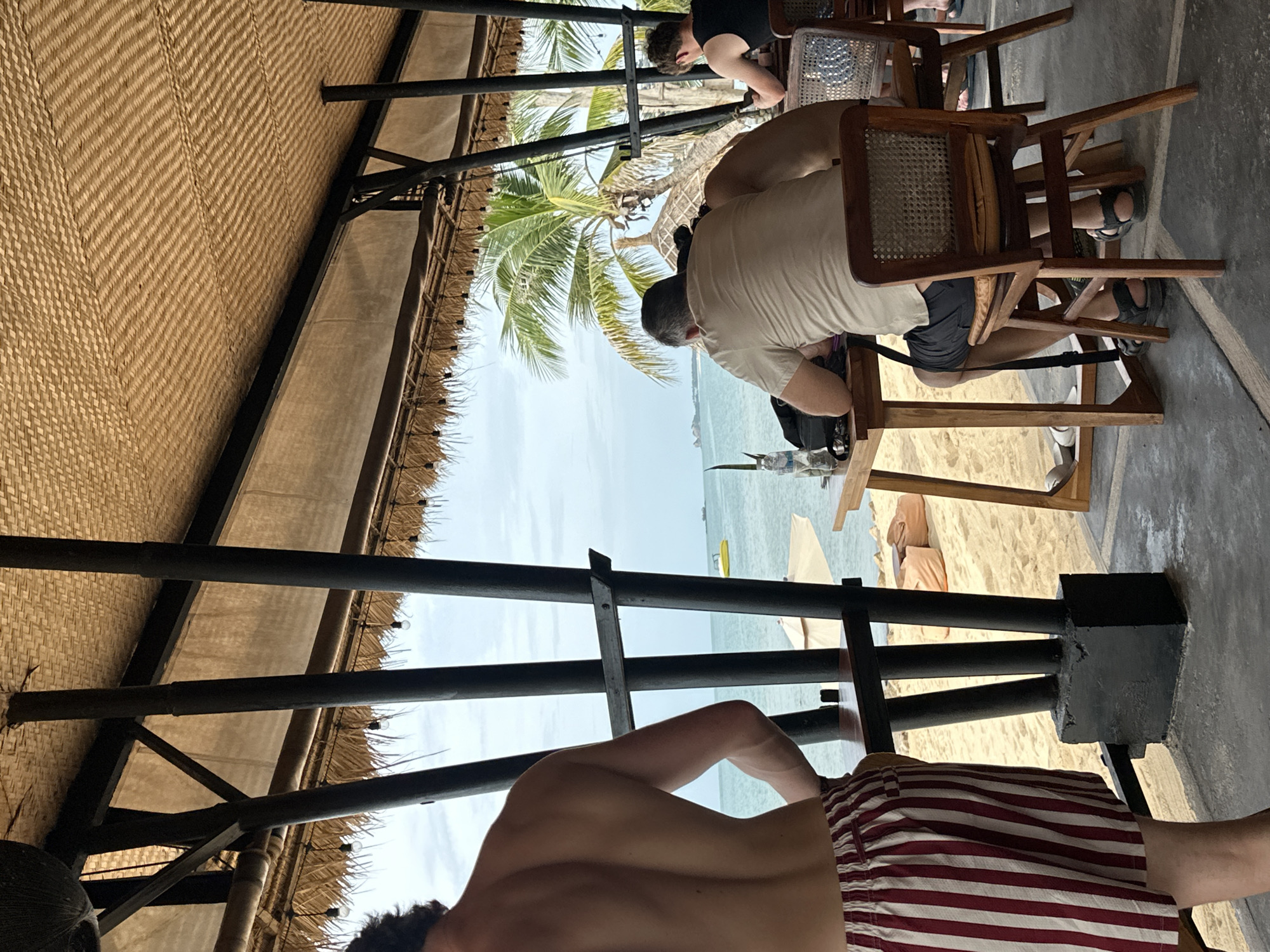 This is a photograph taken outdoors, likely at a beachside restaurant or bar in Ko Samui District, Thailand. The foreground is dominated by a person wearing red and white striped shorts, facing away from the camera. Behind them, a table with a wooden frame and a woven seat is occupied by a person wearing a light-colored t-shirt and dark shorts, looking down at the table. To their right, another person in a black tank top is seated at a similar table, also looking downwards. The environment features a thatched roof overhead supported by dark pillars. Beyond the seating area, a sandy beach with scattered beanbag chairs and umbrellas can be seen, leading to the ocean. Palm trees are visible in the background, suggesting a tropical setting. The sky is overcast.