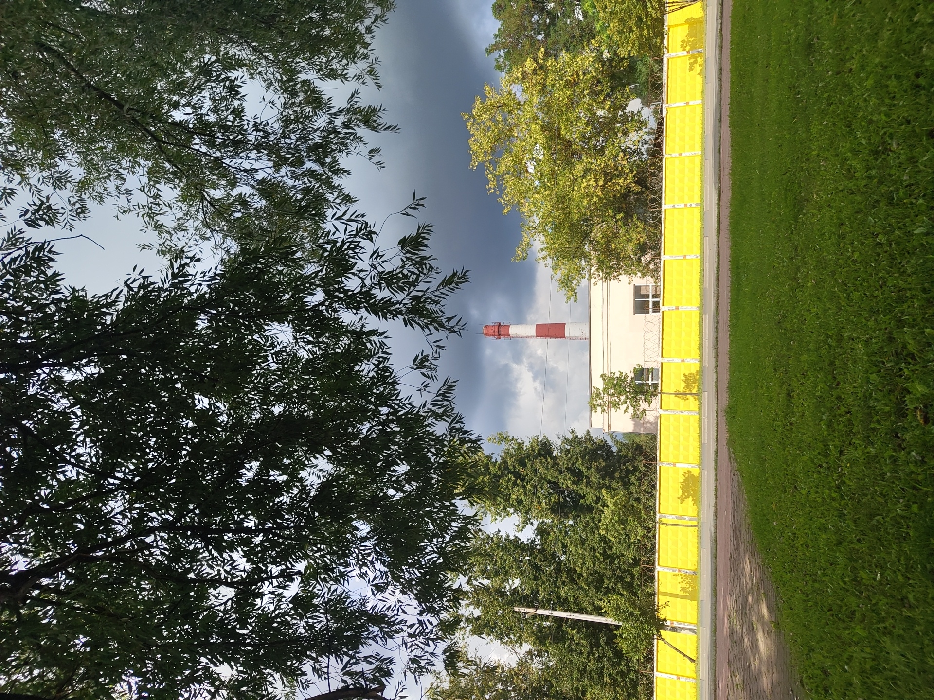 The image depicts an industrial scene in Ramenskoye, Russia, featuring a tall, red and white striped chimney with a building at its base. The building is a light-colored, rectangular structure with windows. A bright yellow concrete fence runs horizontally across the mid-section of the image, topped with barbed wire. Behind the fence, a dense line of green trees stands, with a cluster of lighter green, possibly autumnal, trees to the right. The sky above is a mix of dark, stormy clouds and patches of lighter sky, suggesting an overcast day with potential for rain. In the foreground, a well-maintained strip of green grass is visible, separated from a paved walkway by a darker brick-like surface. Lush green foliage from overhanging trees drapes down from the top of the frame, partially obscuring the sky. No people are visible in the scene.