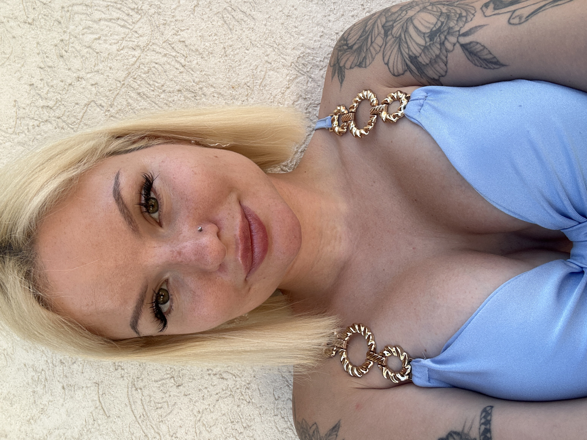 This is a close-up, head-and-shoulders shot of a person with blonde hair and a light blue bikini. The person is lying down, with their head slightly tilted to the side. They have on heavy eye makeup, including dramatic false eyelashes, and a nose ring. Their lips are painted with a matte lipstick. A tattoo is visible on their left shoulder and upper arm, featuring floral and vine elements. The bikini has gold chain detailing on the straps. The background is a textured, light-colored surface, possibly a wall or outdoor seating area. The lighting suggests it might be daytime, with soft, diffused light. The overall mood is relaxed and casual. The image was taken in Budva, Montenegro. There is no visible text in the image.