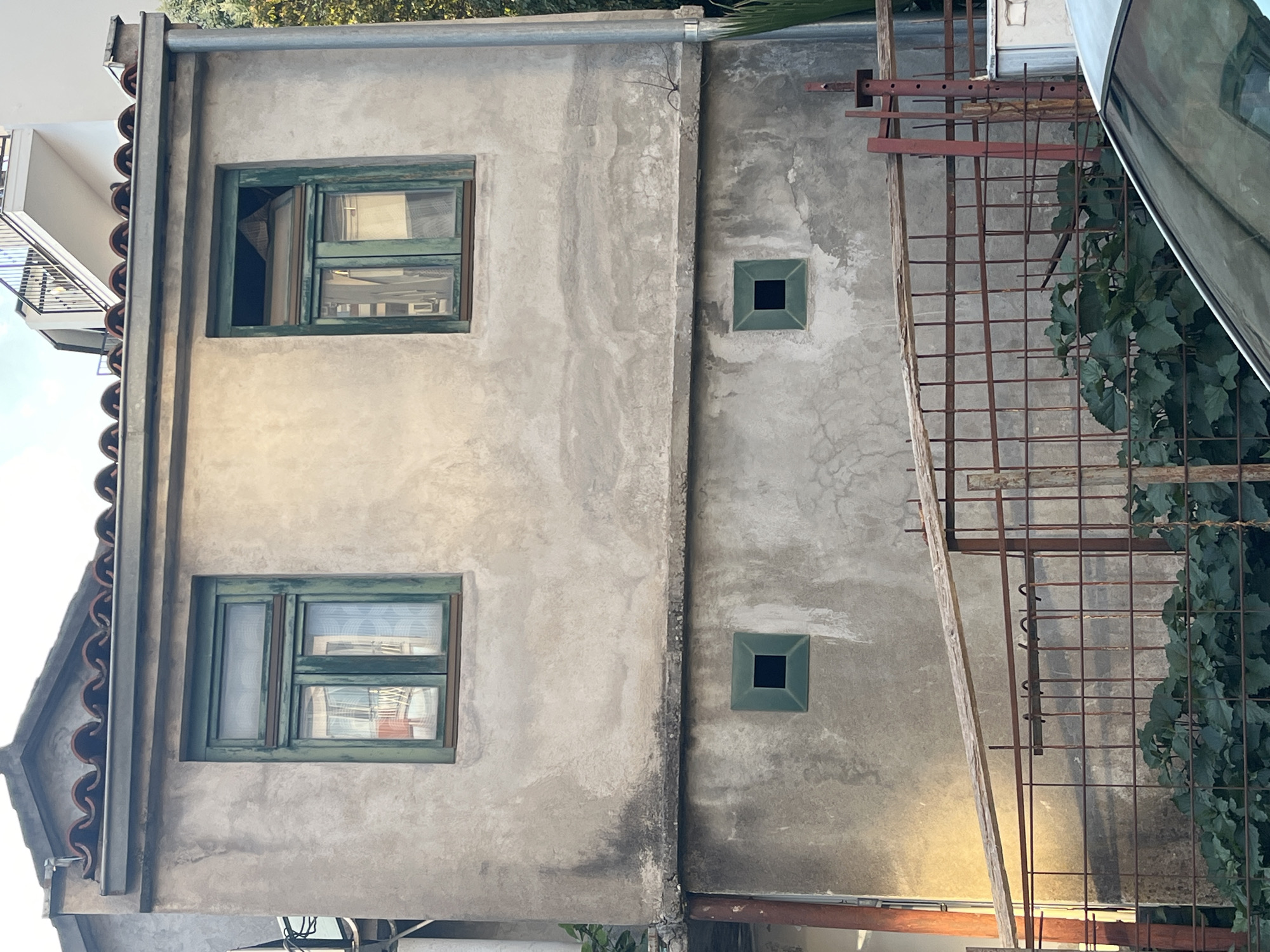 The image shows the exterior of a two-story building in Budva, Montenegro. The building has a weathered, gray concrete facade. The upper story features two green-framed windows, one reflecting an interior scene with curtains and a red structure, and the other with its upper portion open and a triangular object visible inside. The lower story has two small, square, green-framed openings, also appearing to be windows or vents. A horizontal concrete ledge separates the two stories. In the foreground, there is a rusted metal fence with a wooden plank laid across it. Behind the fence, lush green foliage is visible. A portion of a white vehicle is visible in the bottom right corner of the frame. The sky is visible at the top, with some clouds and trees on the right side. The architecture suggests an older, possibly residential, structure.