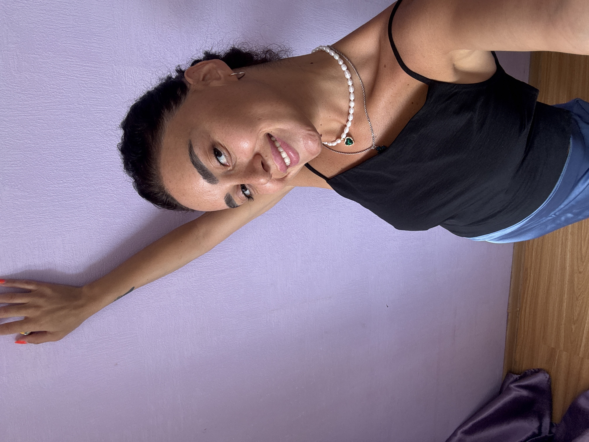 A woman with dark hair pulled back and wearing a black tank top and blue pants poses against a textured purple wall in what appears to be an indoor setting with wooden flooring. She has a necklace made of pearls and a pendant with a green stone, and her nails are painted orange. Her right arm is extended upwards, leaning against the wall. The scene is lit by natural light, casting subtle shadows. The presence of a draped purple fabric on the left suggests a home environment. There is no identifiable information about the location being Tbilisi, Georgia.