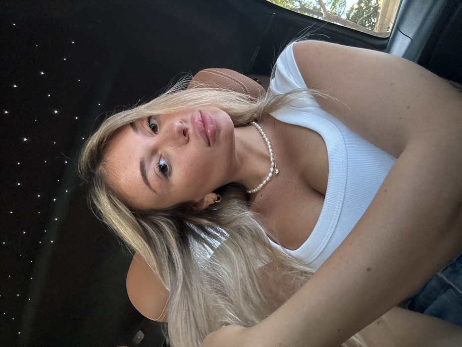 A woman with blonde hair and light skin poses for a selfie inside a vehicle. She wears a white top and a pearl necklace. The vehicle's interior features a dark ceiling with embedded lights mimicking stars. She is angled towards the camera, with her face taking up much of the frame. A brown leather seat is visible in the background. Outside the car window, trees are visible. The image is well-lit, showing the woman's features clearly.