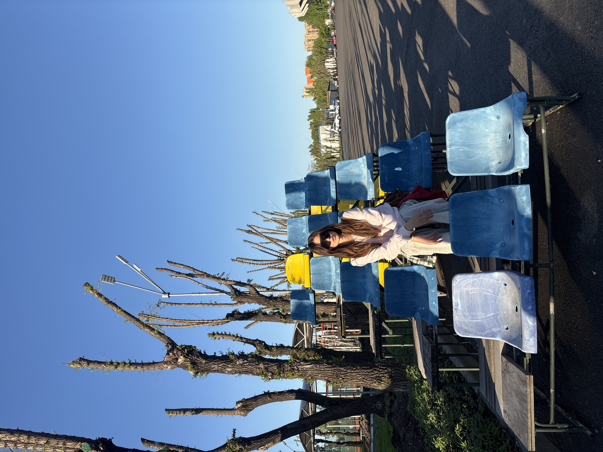 A medium shot depicts a woman with long brown hair, wearing sunglasses, a light pink top, and grey pants, lying on a row of blue stadium seats, with a yellow seat visible. The location is Krasnoyarsk, Russia. A large tree, recently pruned with budding green leaves, partially obscures the stadium seating. The background includes a dark asphalt surface with tree shadow patterns and distant buildings under a clear blue sky. The photo appears to be taken during the day.