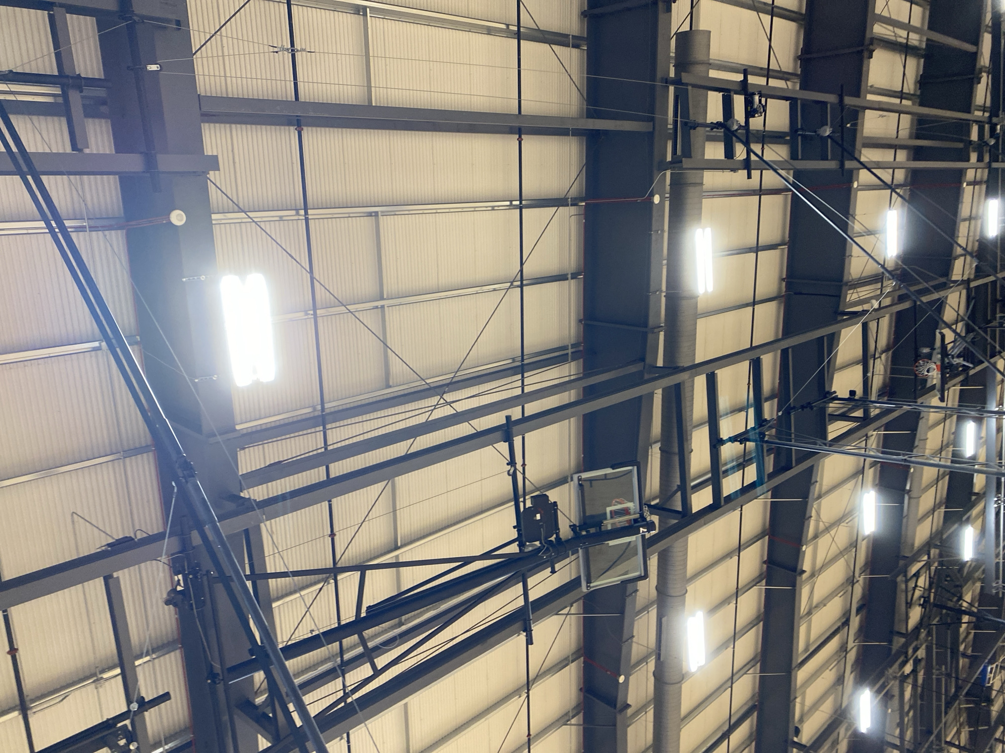 This image shows the interior of a gymnasium, looking up at the ceiling structure. The ceiling is composed of corrugated metal panels supported by dark gray steel beams and trusses. There are fluorescent lights mounted on the ceiling, some of which are illuminated, casting a bright glow. Visible on the structure are various pipes and conduits, as well as what appear to be basketball hoops and backboards suspended from the ceiling. The overall scene suggests an indoor sports facility. The location in Virginia Beach, United States of America is not discernible from the image itself.
