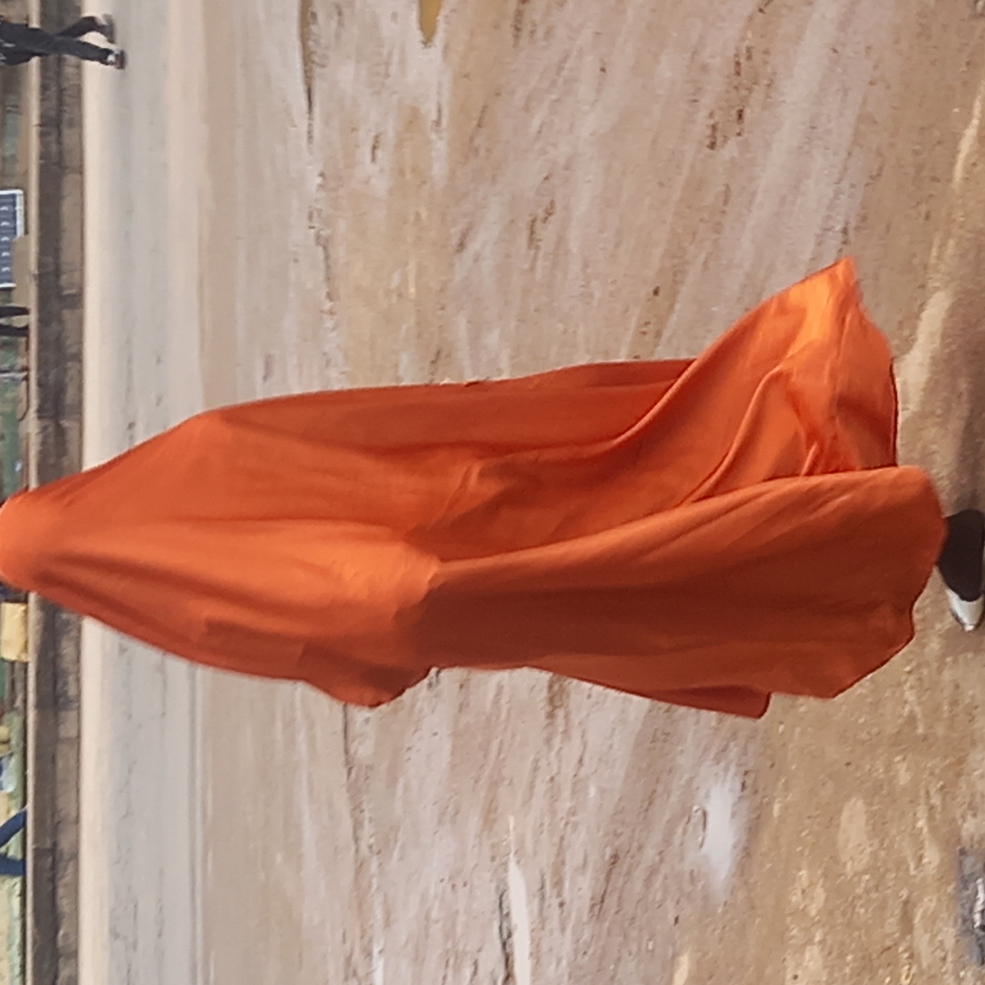 A street in Dadi Riba, Nigeria is the setting for this photo. An orange cloth billows or drapes over an object or person, obscuring it from full view. The ground surface appears to be a mix of paved road and dirt. A person wearing black and white shoes is seen at the right edge of the frame. In the background, on the left, glimpses of figures and structures are visible, suggesting a populated area.