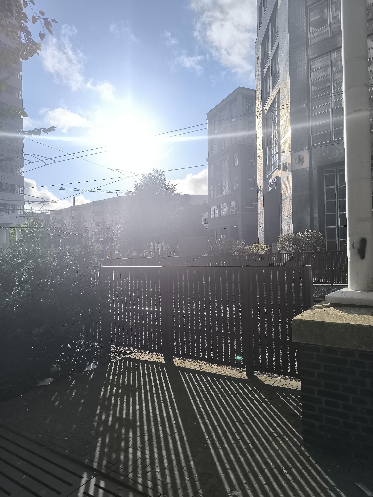 The photograph captures a sunny outdoor scene in front of modern buildings, possibly in a European city. The sun is bright, creating lens flare and casting distinct striped shadows on the ground from a wooden fence. The buildings appear to be a mix of brick and glass, with multiple stories and windows. In the foreground, a dark wooden fence runs horizontally across the frame. Beyond the fence, trees and bushes are visible. Overhead, power lines and a construction crane are present, suggesting an urban or developing area. The sky is blue with some scattered clouds. The lighting indicates it is daytime, likely mid-morning or afternoon. The scene is devoid of any people.