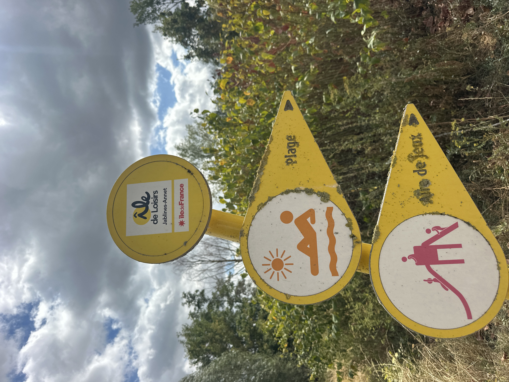 The image shows a yellow directional signpost located in Jablines, France, specifically at the "Île de Loisirs Jablines-Annet." The signpost has three circular signs mounted vertically on a pole. The top sign features the logo for "Île de Loisirs" and the text "Jablines-Annet" along with the "Île de France" emblem. Below this, the second sign displays an orange pictogram of a person sunbathing on a beach, with a sun above and waves below. A yellow arrow pointing right from this sign has the word "Plage" (Beach) written on it. The third sign, at the bottom, shows a pink pictogram of children playing on a slide. A yellow arrow pointing right from this sign has the text "Aire de Jeux" (Play Area) written on it. The background consists of lush green trees and foliage under a cloudy sky. The signposts are weathered, with some signs of moss and discoloration.