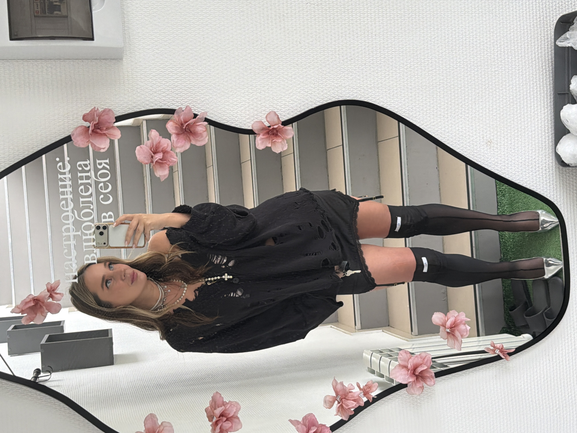 A young woman is captured in a mirror selfie, with the original image rotated horizontally. She has long, light brown hair and blue eyes, and is looking slightly upwards. She is dressed in a black, distressed mini-dress or tunic, black sheer thigh-high stockings with white accents, and silver pointed-toe high heels. She wears multiple layered necklaces, one with a cross charm, and holds a light-colored smartphone (likely an iPhone) to take the picture.
The mirror has an irregular, wavy black frame, which is decorated with numerous delicate pink artificial flowers. The reflection in the mirror shows an indoor setting, featuring light-colored stairs and walls, along with a patch of green flooring or turf near her feet.
Outside the mirror's frame, a white textured wall is visible, along with several grey rectangular containers and a black tray holding white, soft objects (possibly cotton balls).
Reflected in the mirror, upside down and backwards, is the Russian text "настроение: влюблена в себя," translating to "mood: in love with myself." The word "проблема" (problem) is also partially visible near this phrase. The scene appears to be indoors, likely in Chaikovsky, Russia, conveying a fashionable and self-confident aesthetic.