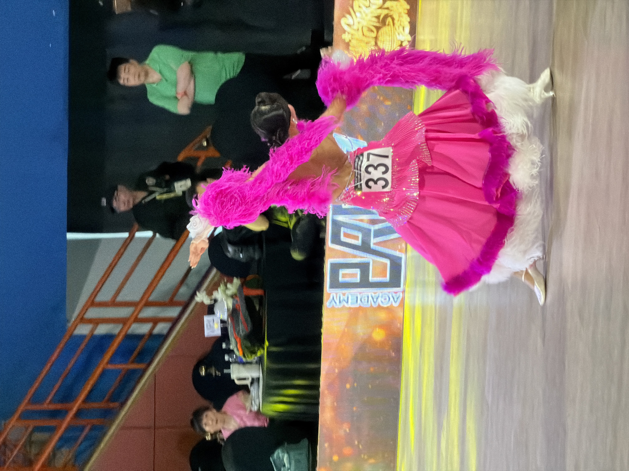A young person in a vibrant pink dance costume with feather accents is performing on a polished floor in what appears to be an indoor sports arena or ballroom. The costume features a full skirt, embellished with sequins, and white furry trim at the hem. The dancer has their back to the camera, showcasing the elaborate design of the outfit. A bib with the number "337" is pinned to their back. In the background, there's a stage backdrop with "ACADEMY" and "EA" visible, illuminated by yellow lighting. Spectators and event staff can be seen in the bleachers and behind the performance area, including a woman in a pink shirt, two men in dark shirts and caps, and a man in a green polo shirt. The environment suggests a dance competition or performance event. The specific location in Hanoi, Vietnam, cannot be determined from the visual information provided.
