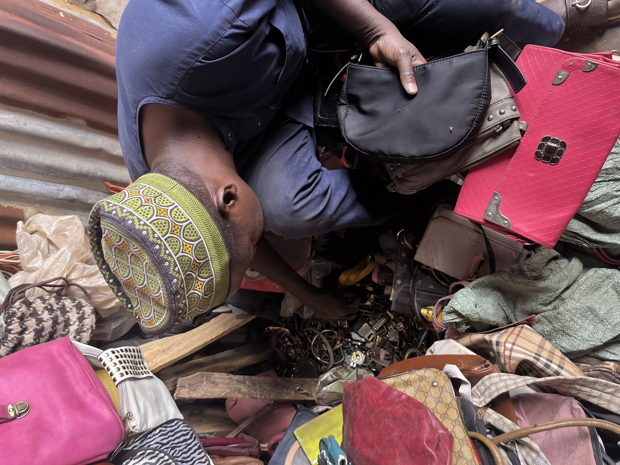 A man in a blue top and a patterned green and brown hat is kneeling and searching through a pile of bags, jewelry, and other items. He is holding a black purse in his right hand. The scene is cramped and filled with various objects. The location is Jos, Nigeria.