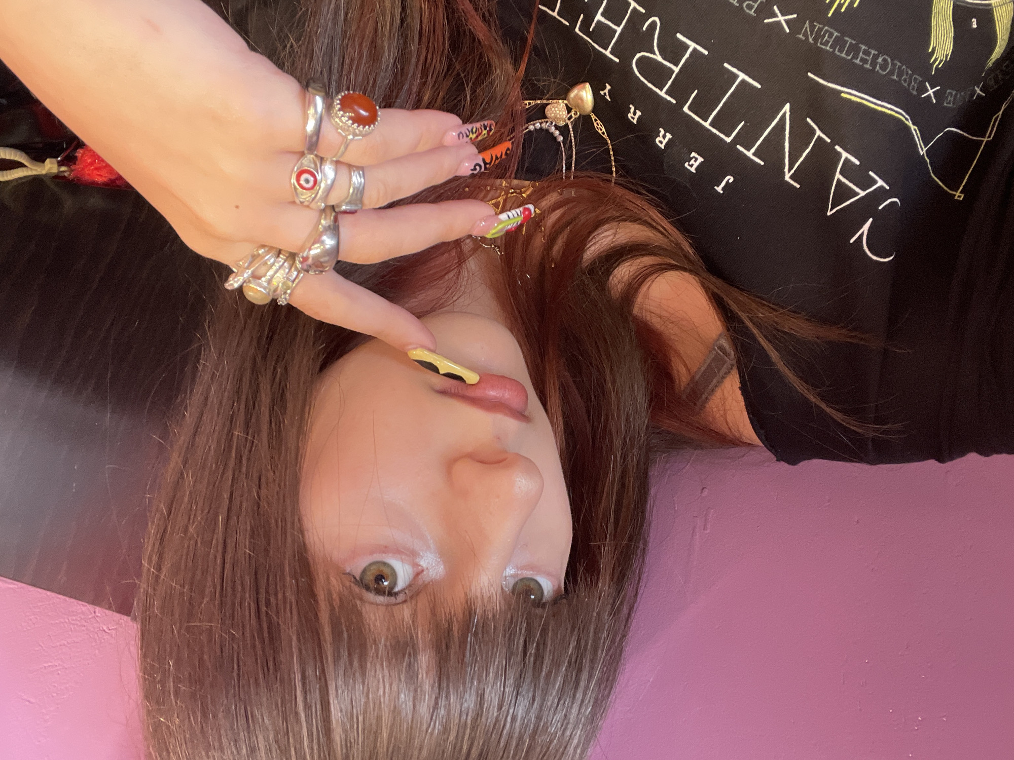 A woman is photographed from an inverted perspective. She lies on a pink surface, wearing a black t-shirt with the white text "CANTRELL JERRY x BRIGHTEN x". Her hair is brown with bangs covering her forehead. She has light skin and light brown eyes. She is wearing multiple silver rings on her fingers, and her nails are decorated with colorful designs. There is a table behind her to the left. The location is Raanana, Israel.