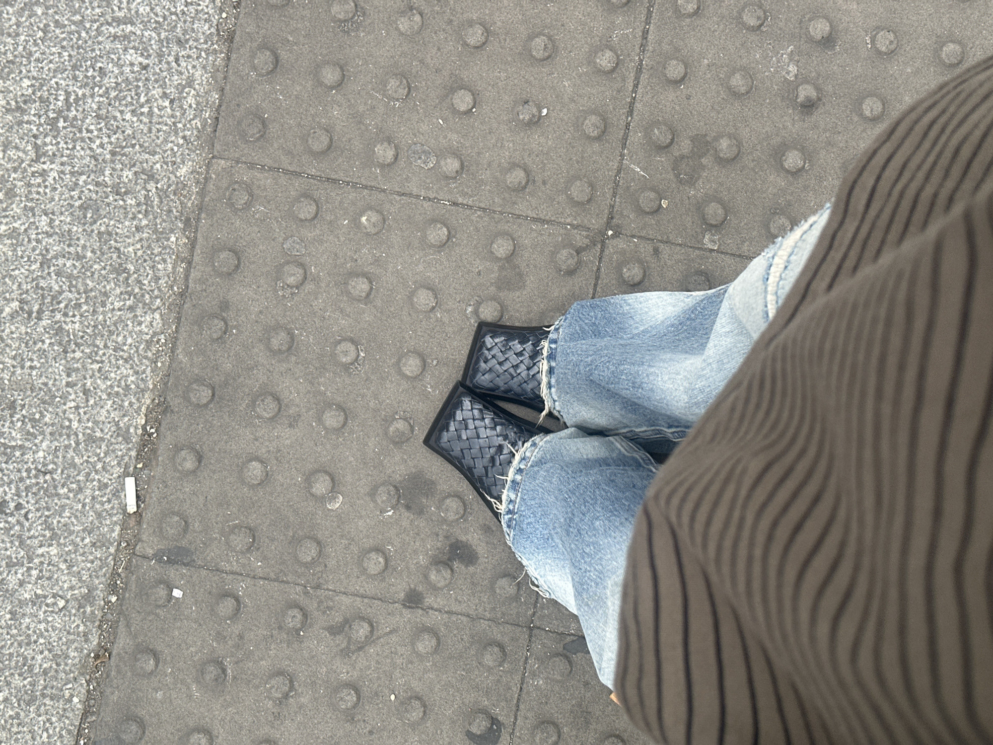 An overhead shot shows a person standing on a gray concrete sidewalk. The sidewalk is textured with rows of raised circular bumps. The person is wearing light blue denim jeans with frayed hems and black, woven leather shoes. A portion of the person's striped, brown sweater or coat is visible on the right side of the frame. The location is London, United Kingdom.