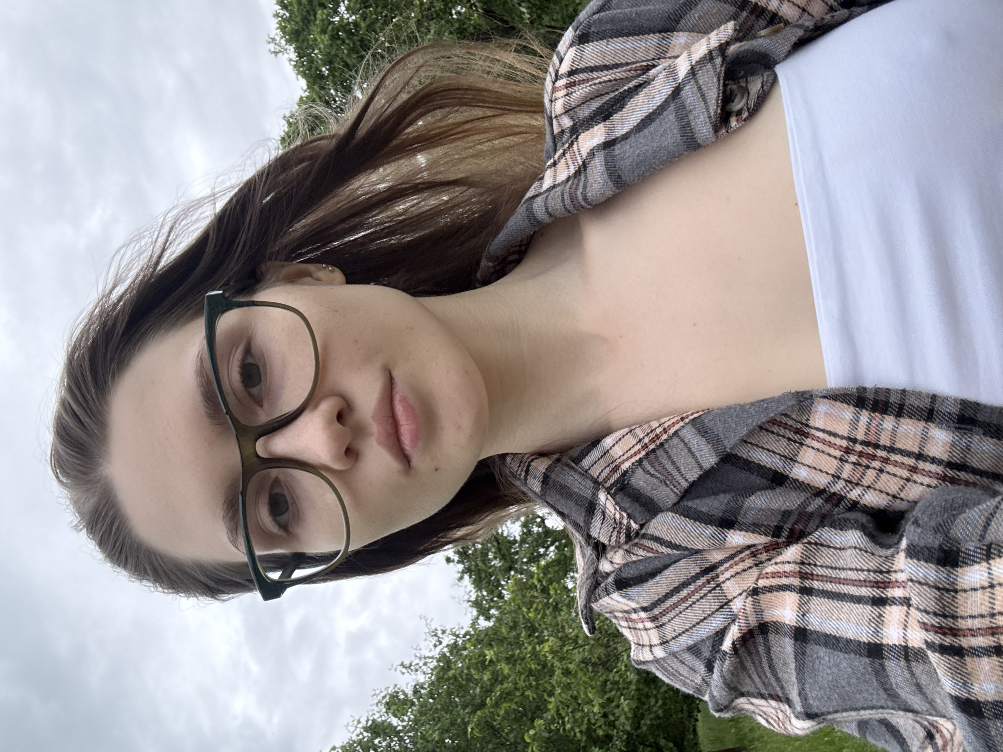 A young woman with light brown hair and glasses is taking a selfie outdoors. She is wearing a plaid shirt over a white top. The background shows trees and an overcast sky. There is no discernible information about the location in Saint Petersburg, Russia, or any specific actions or interactions. The scene is a straightforward portrait.