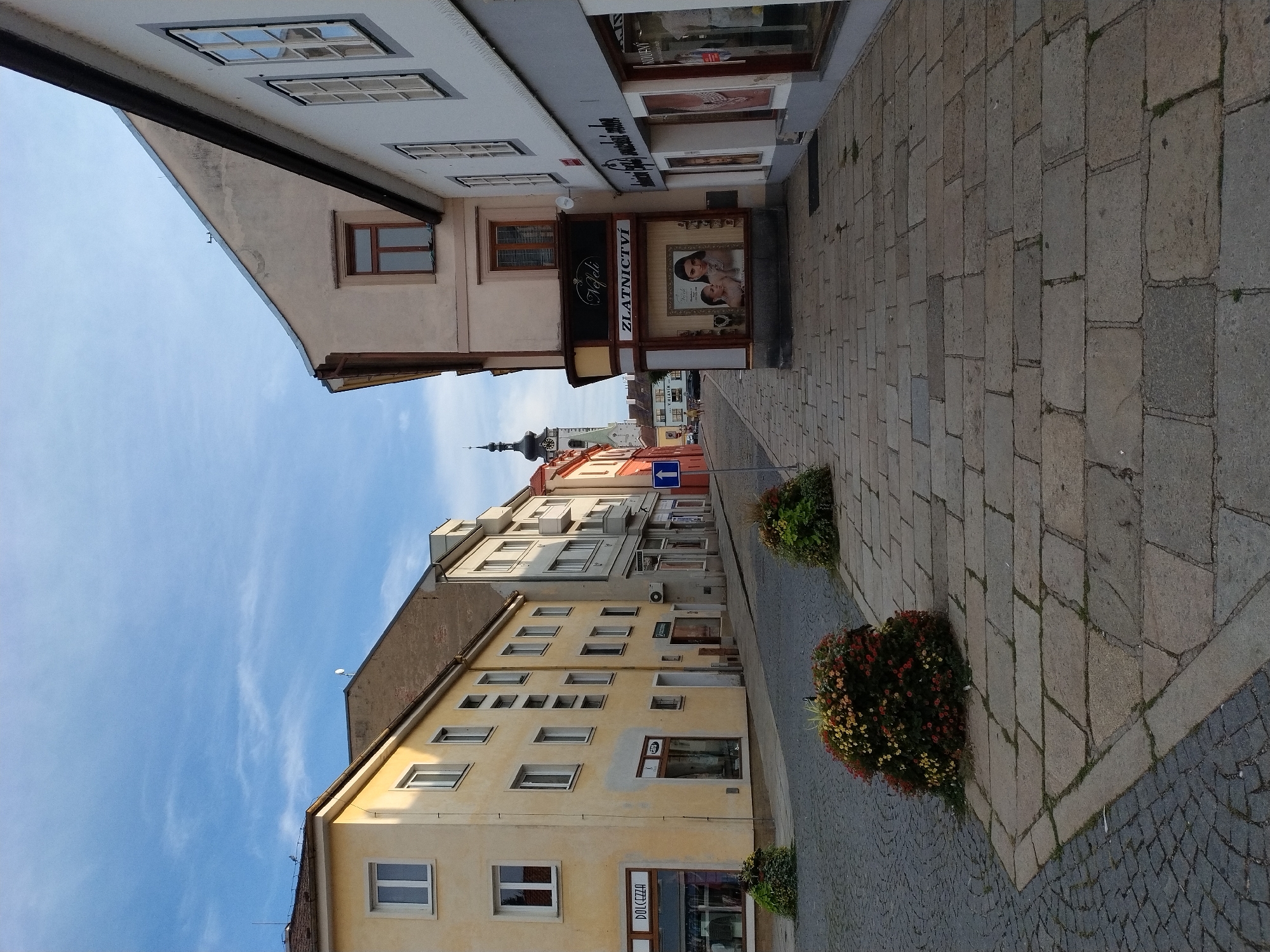 The image depicts a street scene in Písek, Czechia, featuring a cobblestone pavement and buildings on either side. In the foreground, a large planter with blooming flowers sits on the right side of the sidewalk. The sidewalk itself is paved with large, irregular stone slabs. To the left, the pavement transitions to smaller, more uniform cobblestones.
On the left side of the street, a yellow building displays several windows and a storefront with signage reading "DOLCEZZA". Further down, another storefront has signage that is partially obscured.
On the right side of the street, a building with a beige facade has a prominent storefront for a jewelry store, indicated by the sign "ZLATNICTVÍ" and images of jewelry and models in the window. Above this storefront is a sign that reads "Nefeli". Adjacent to this building is another white building with windows and storefronts, one of which has the partially visible sign "KOUPNY".
In the background, a church tower with a dark spire rises above the buildings. The sky is blue with scattered white clouds. The overall impression is a charming, historic street in a European town.