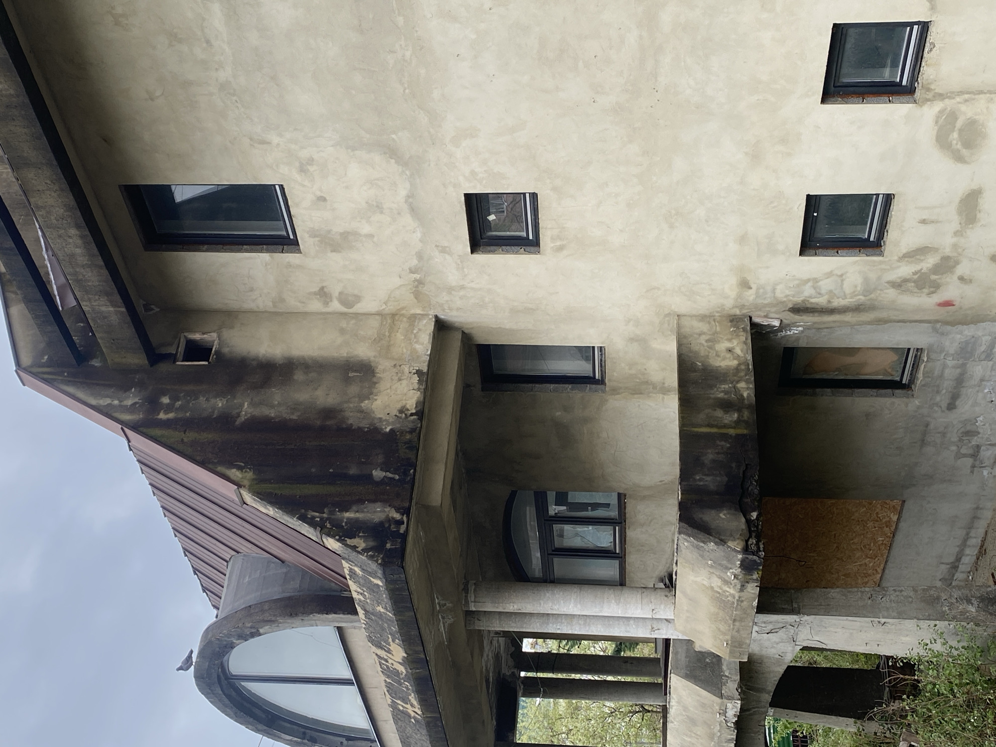 The image displays a multi-story concrete building in Sochi, Russia, viewed from a low angle against an overcast sky. The architecture is characterized by raw, exposed concrete surfaces, which show signs of wear, discolouration, and possible dampness, suggesting an aged, unfinished, or semi-abandoned state.
Numerous dark-framed windows are scattered across the facade. On the upper levels, there are several small, square windows and a larger rectangular one, some reflecting the grey sky. Lower down, the structure features recessed areas and prominent concrete beams and round pillars, forming what appears to be a covered outdoor space or balcony. Within this area, a large, distinctive curved window is visible towards the bottom left, with a pigeon perched on its frame. Another window opening is boarded up with a light brown plywood sheet on the mid-right side of the image.
A rusty-red corrugated metal roof is visible on the left side, angling downwards from the main concrete structure. Green foliage from trees or bushes can be seen at the bottom right, indicating an outdoor setting. The diffused light suggests it is daytime with cloudy weather. No discernible text is visible on the building.