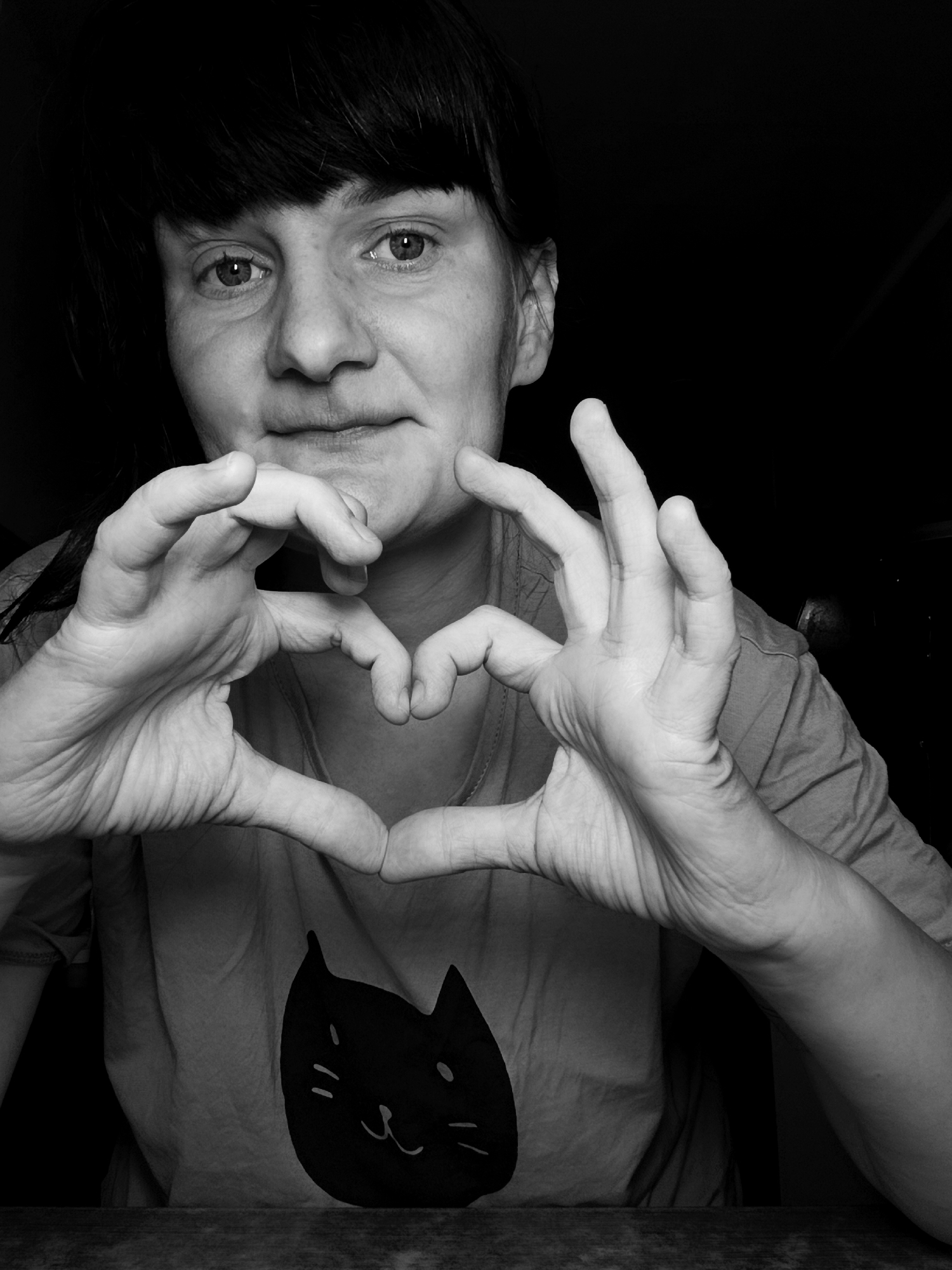 A black and white close-up shows a person with dark hair and a t-shirt with a cat cartoon printed on it, making a heart shape with their hands. The location is Unknown, Unknown.
