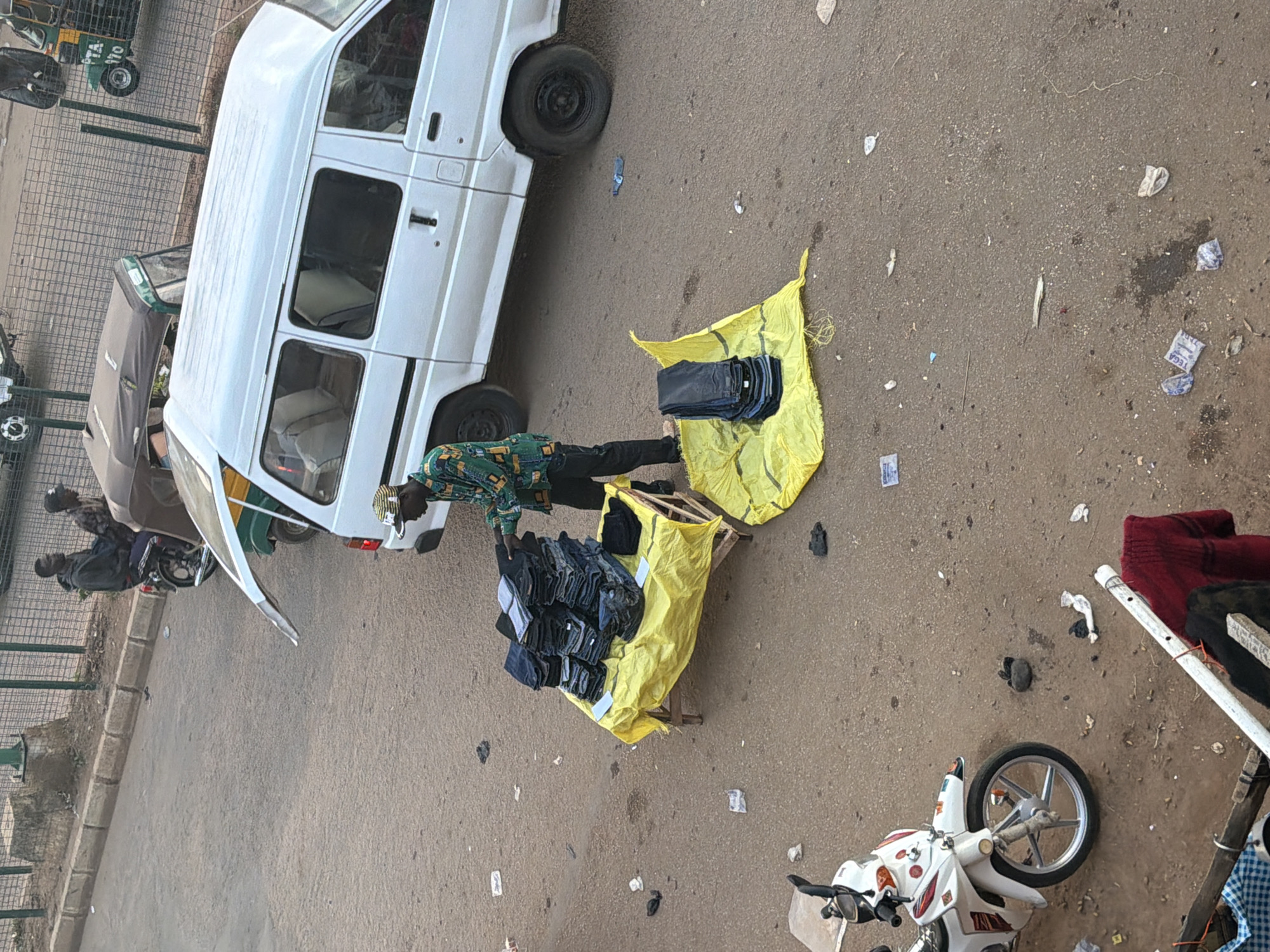 In the city of Bukuru, Nigeria, a street vendor is seen arranging a stack of folded dark-colored pants on a yellow tarp spread over a wooden frame. The vendor, wearing a patterned long-sleeved shirt and dark pants, is in the process of displaying his merchandise. Another smaller stack of pants is placed on a separate yellow tarp on the ground. To the right, a white van with its rear door open is parked on the asphalt road. Further back, two individuals are seated on a motorcycle, and a three-wheeled motorized vehicle is visible. A white motorcycle is parked on the left side of the image. The ground is covered in asphalt with scattered debris.