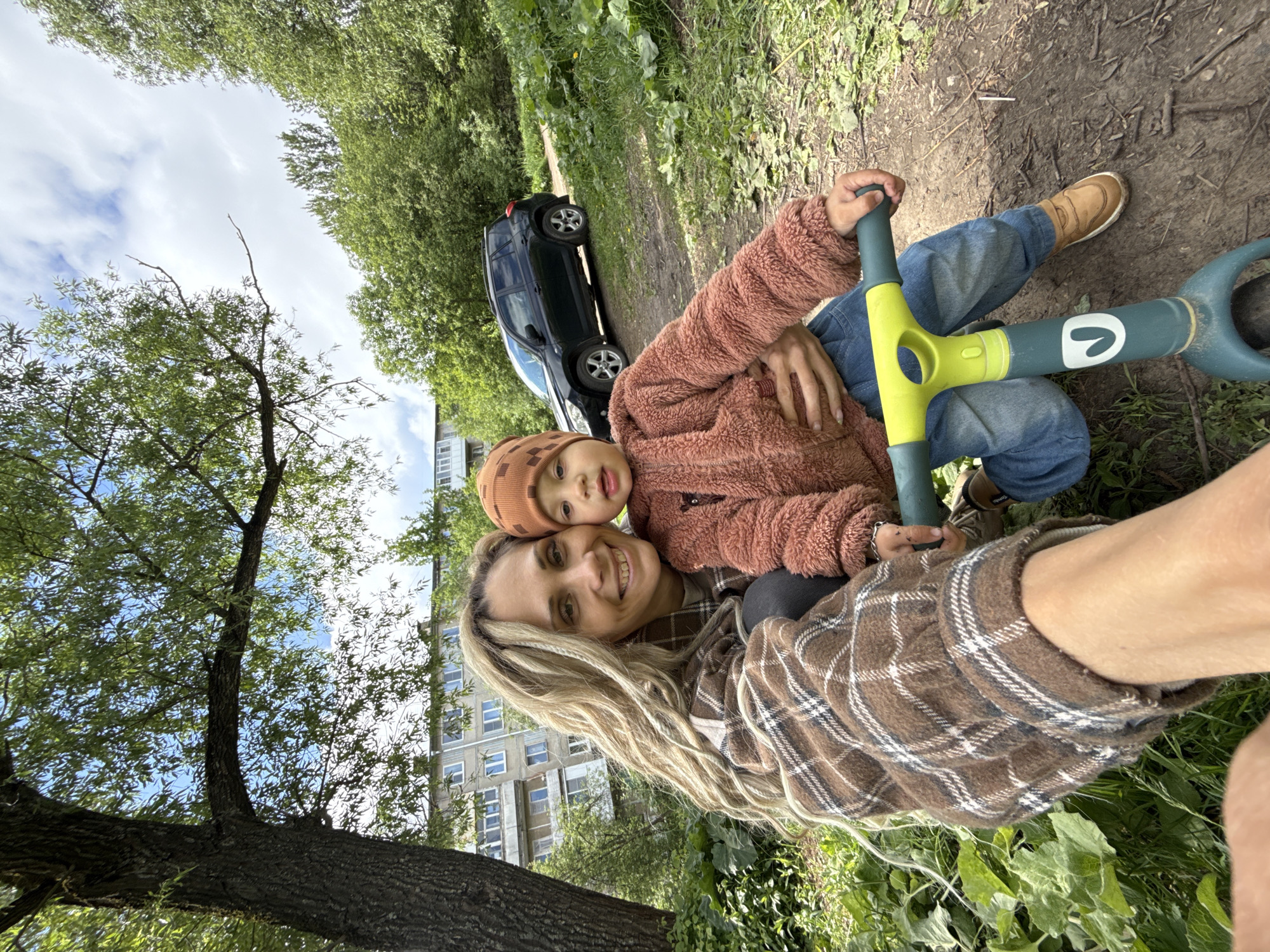 A selfie-style image captures a woman and a young child outside. The woman has long blonde hair and is wearing a brown and white plaid shirt. The child, wearing a brown hat and pink furry jacket, is sitting on the woman's lap. They are both smiling. The child is holding the handlebars of a green and yellow scooter. The backdrop includes a dirt path, greenery, a black car, and an apartment building. The scene appears to be Rusanovo, Russia.