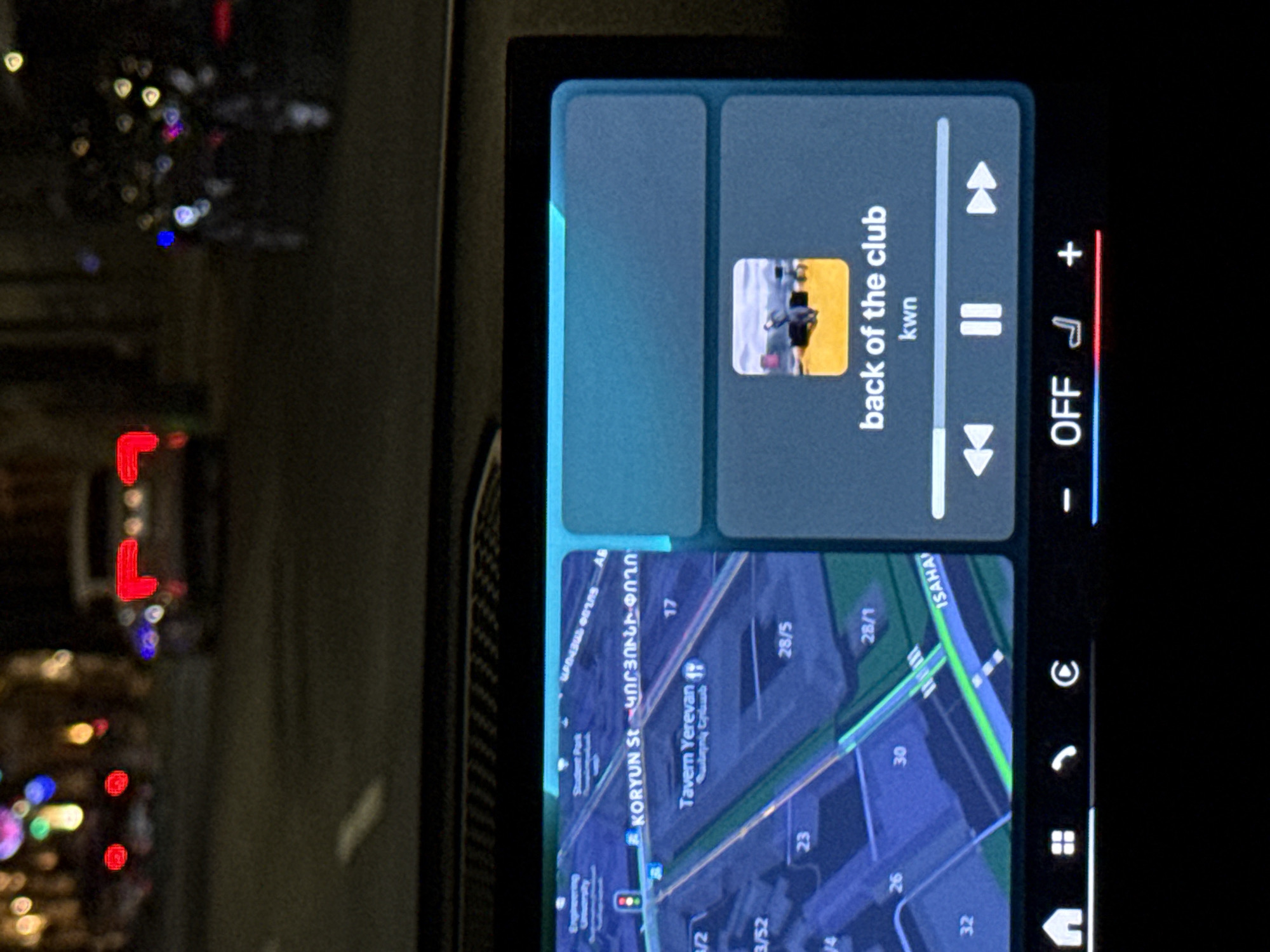 The image shows a car's infotainment screen displaying a map and music player. The map shows Koryun Street in Yerevan, Armenia, near Tavern Yerevan. The music player indicates the song "back of the club" by kwn is playing. The background outside the car window is blurry and out of focus, suggesting it's nighttime.